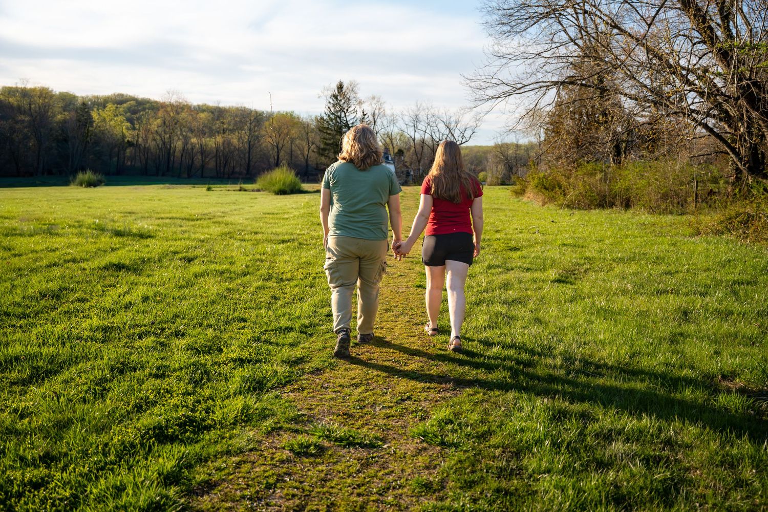 A couple walking hand-in-hand across a green meadow toward a tree line on a sunny day. In Jerusalem mill in Fallston Maryland