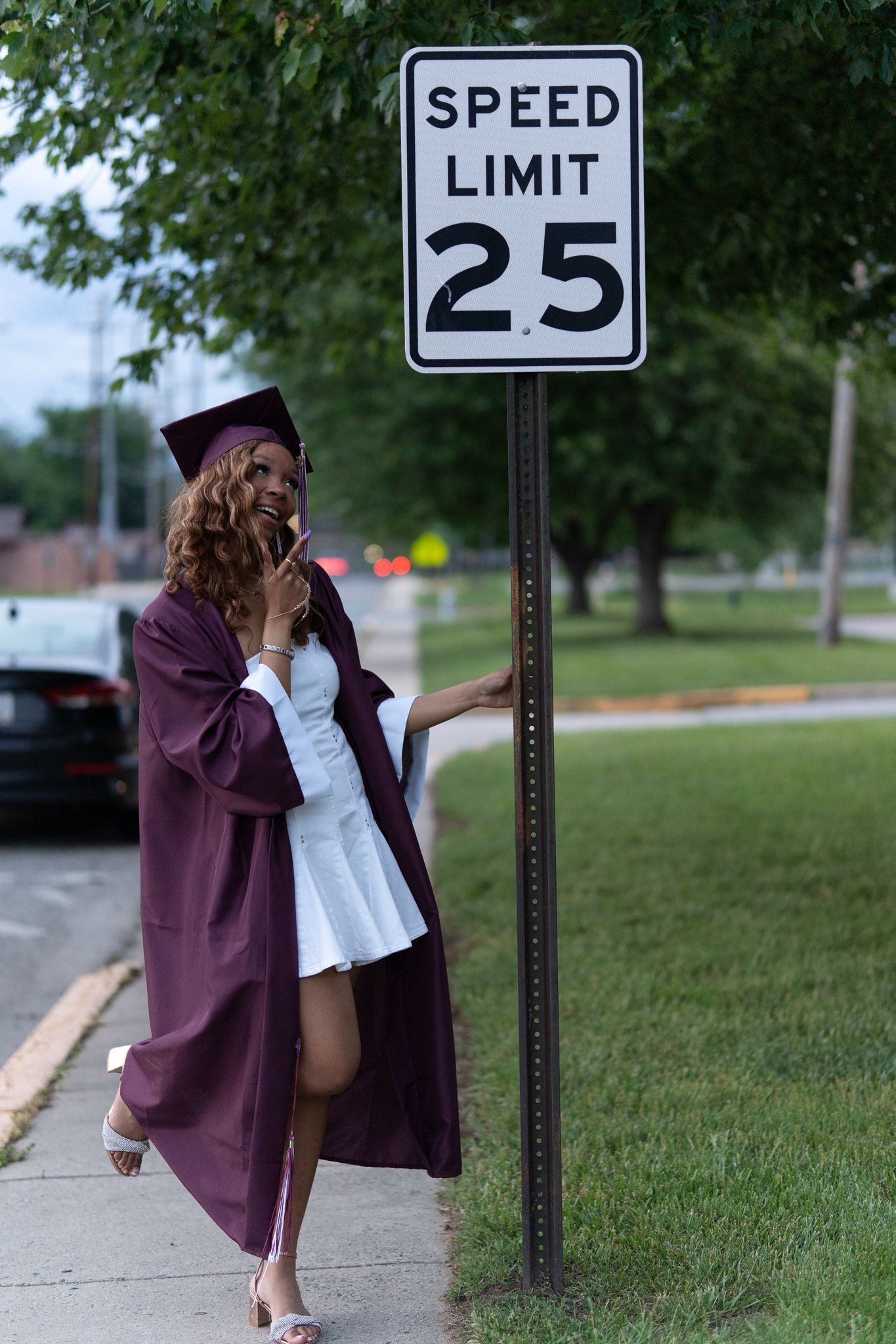 Graduate in maroon cap and gown leaning against speed limit 25 sign, smiling on a sidewalk. In Havre de Grace