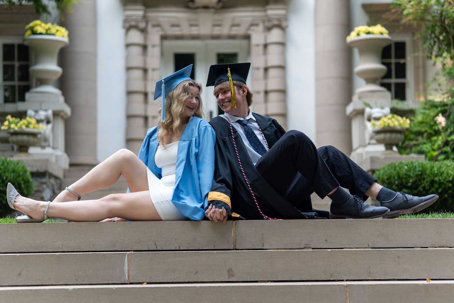 Two graduates in blue caps and gowns sit on steps, smiling and relaxed in an outdoor campus setting. In Bel Air Maryland