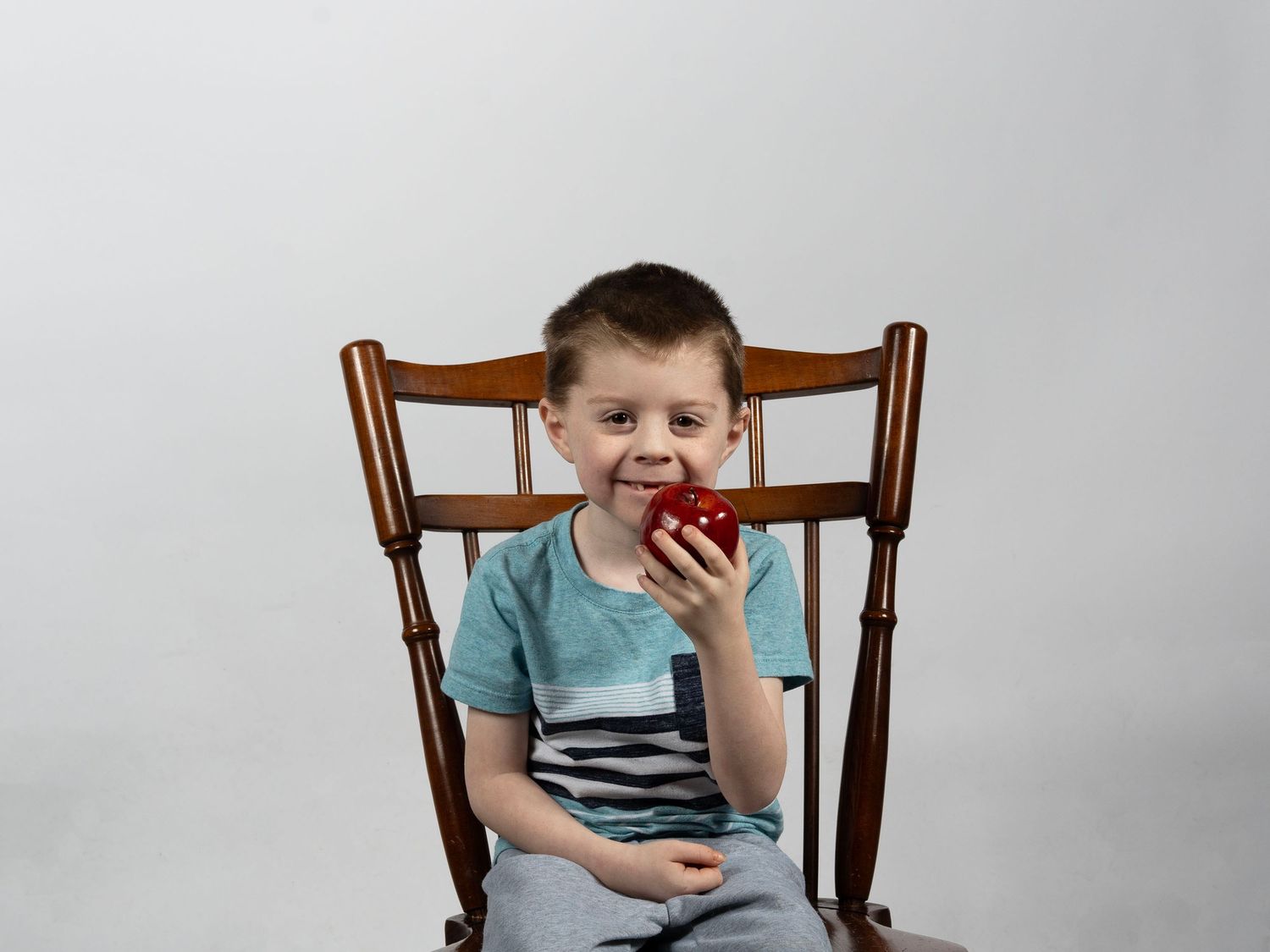 Smiling young boy sits in wooden rocking chair holding red apple against white background. In Bel Air Maryland