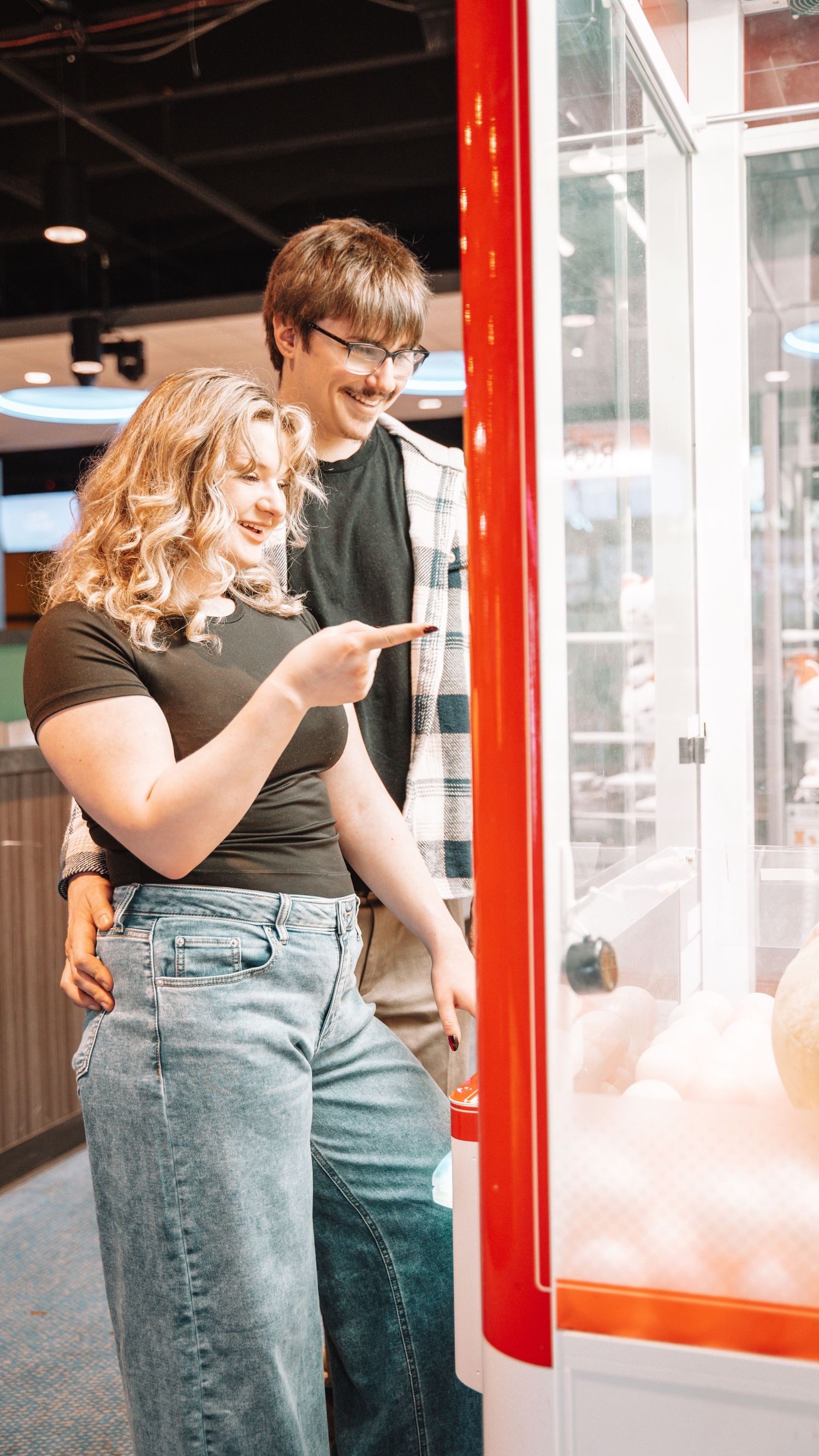 Blonde woman points at claw machine prize while smiling man stands beside her in bright arcade. in Towson Maryland
