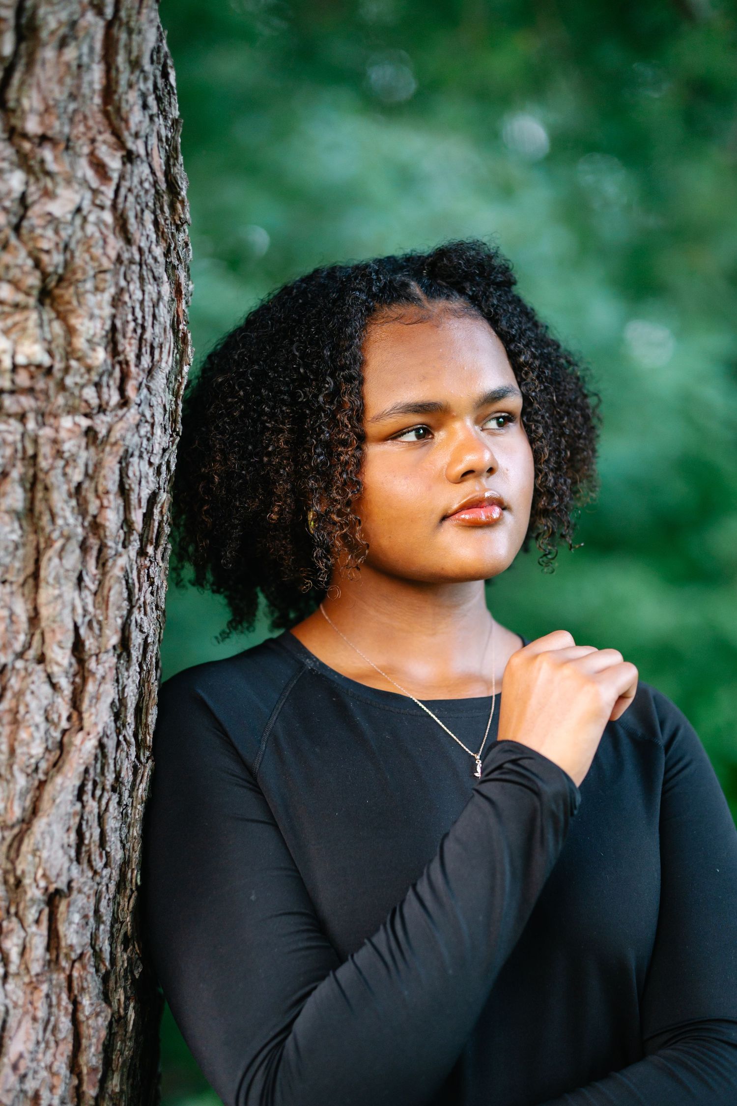 Young Black woman with curly hair in black long-sleeve top leans against tree trunk in lush green park.