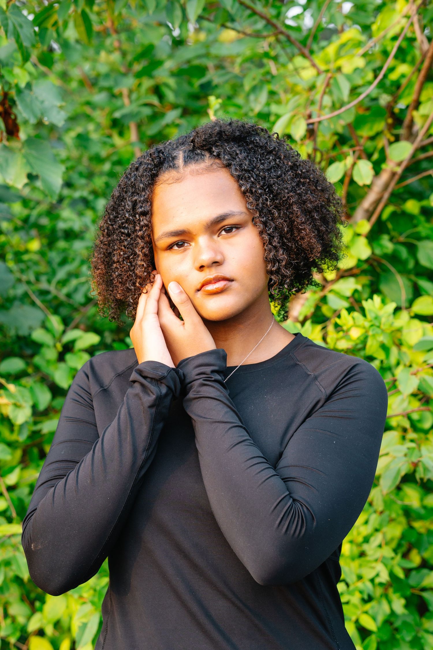 Young person with curly hair wearing black long-sleeve top, posing outdoors with hands on face near green foliage.