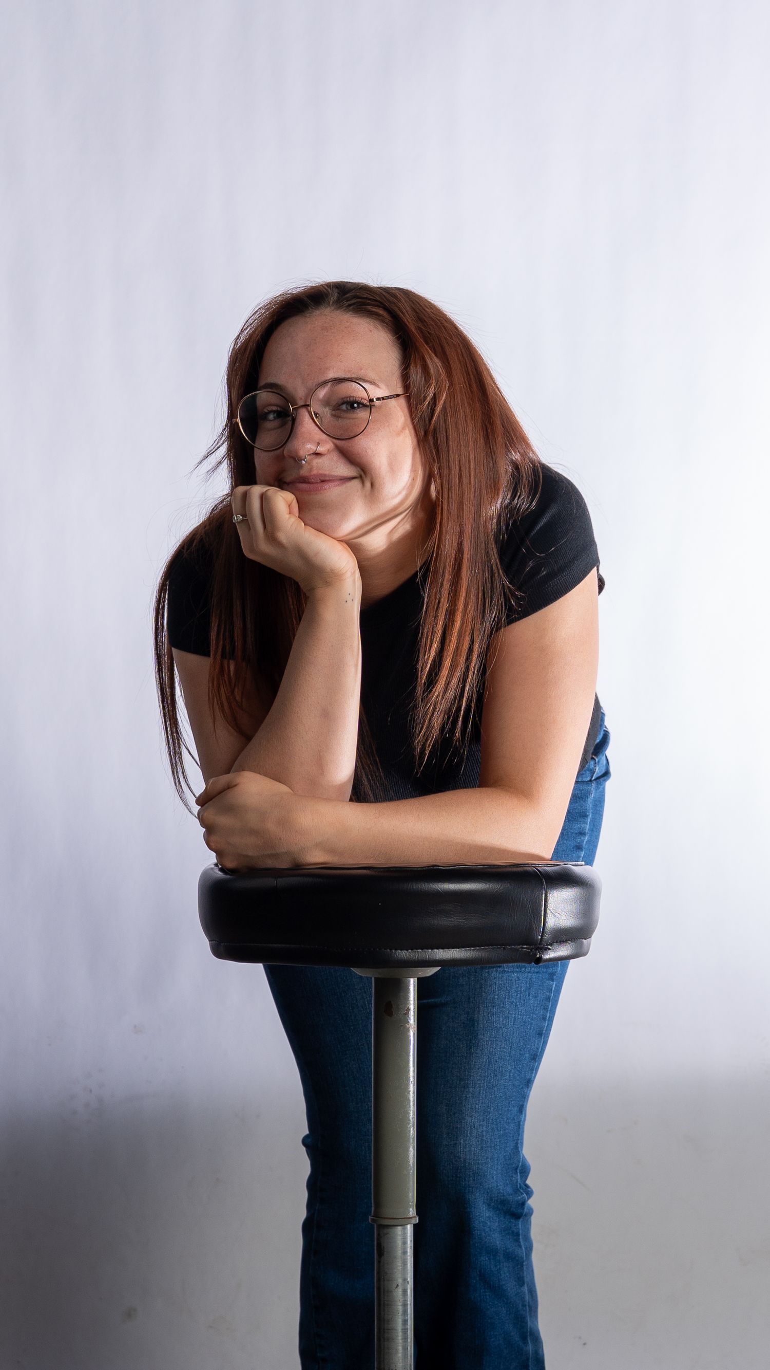 Smiling redhead woman with glasses leaning on a barstool against a white studio background. In Bel Air Maryland
