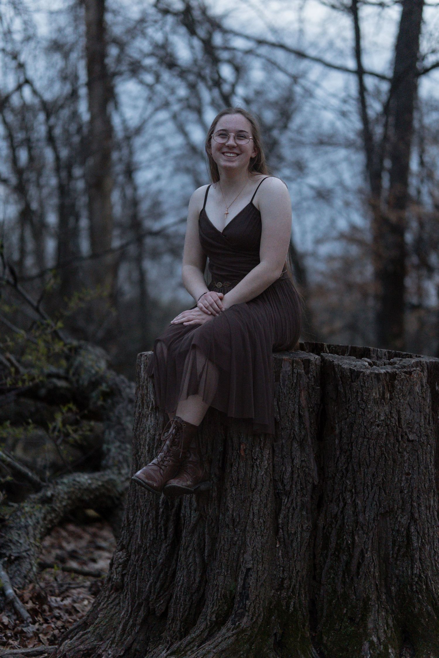Young woman in brown dress and boots sitting on a large tree stump in a forest, smiling at camera. in Maryland Rock state park