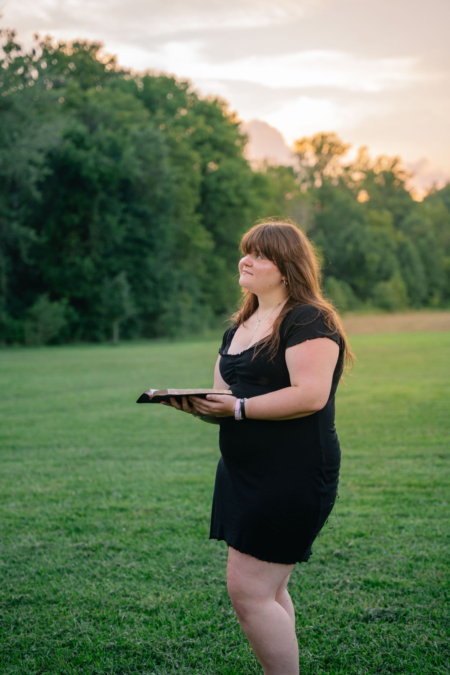 Woman in black dress holding a book outdoors in a green field at sunset.