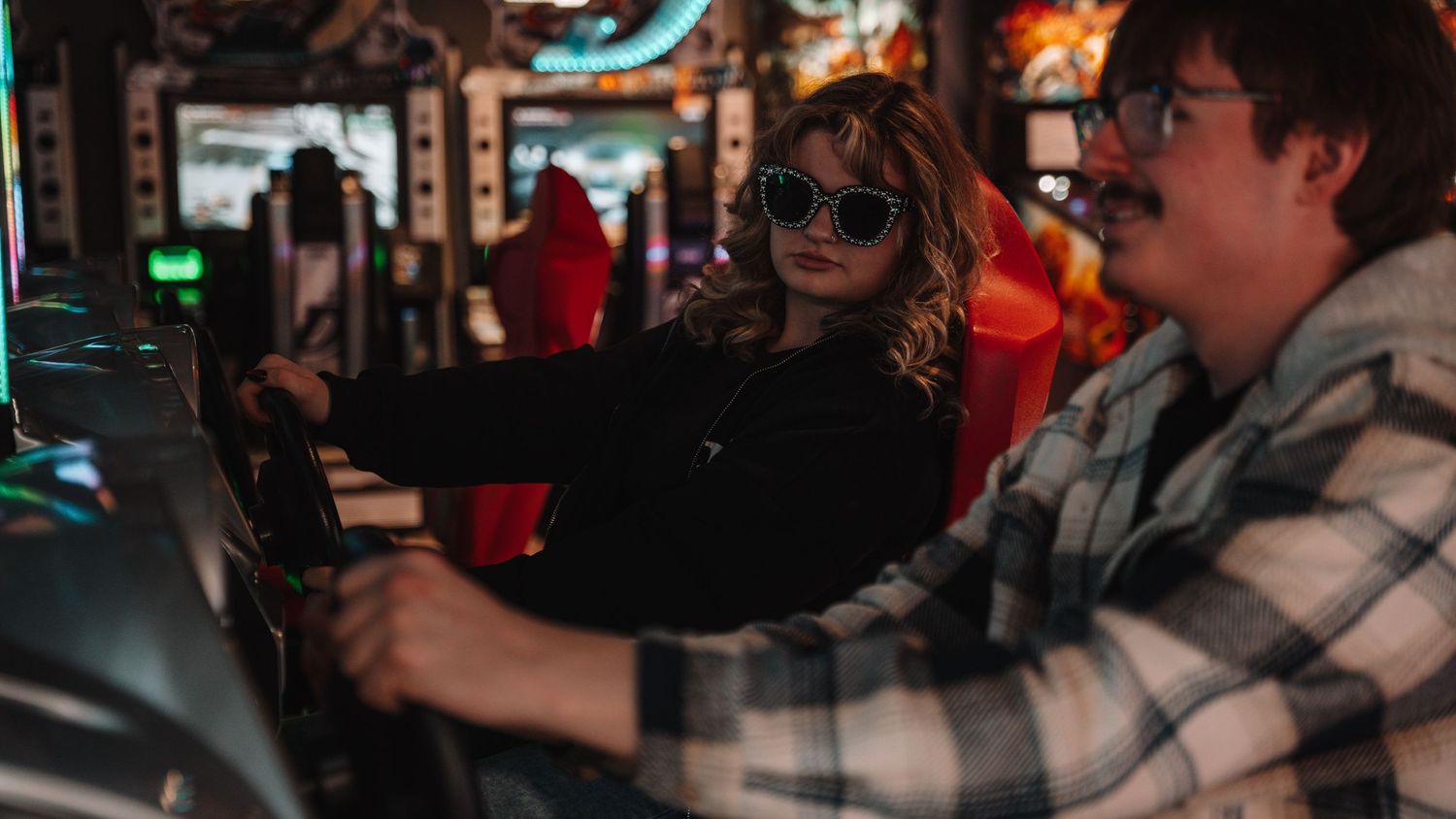 Two young adults playing arcade racing games in a dark gaming arcade, woman wearing sunglasses, man in plaid shirt. in Towson Maryland