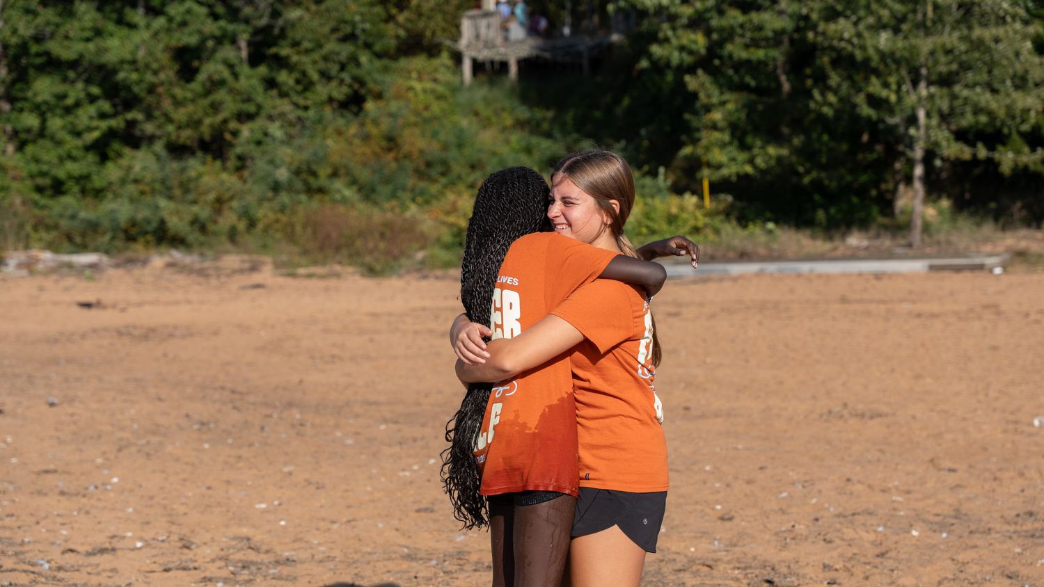 Two women in orange shirts share a warm hug on a sandy beach with trees in the background.