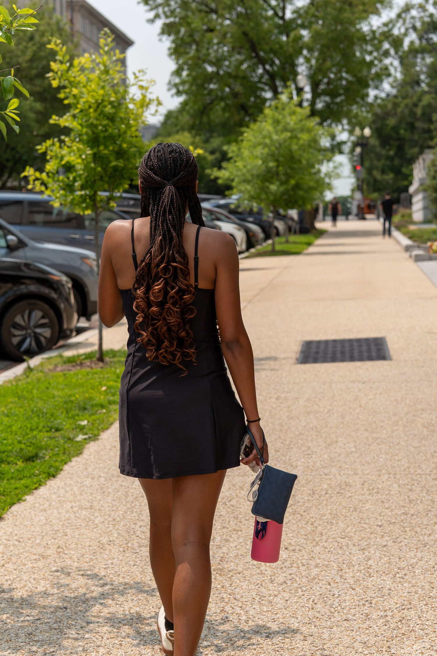 Young Black woman with braided hair walks down a sunny sidewalk in a black dress, holding a pink bottle and small bag. In Washington DC