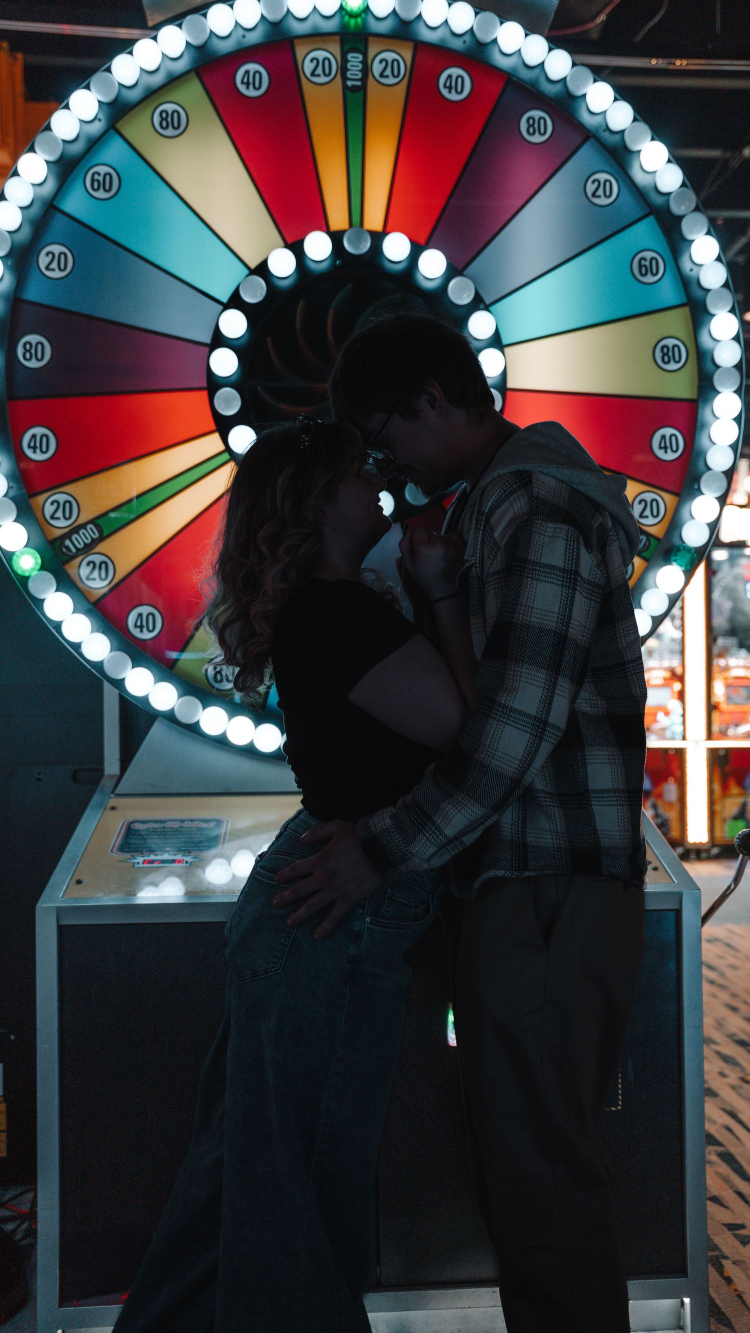 Couple sharing a kiss in front of a illuminated spinning prize wheel at an arcade. In Towson Maryland