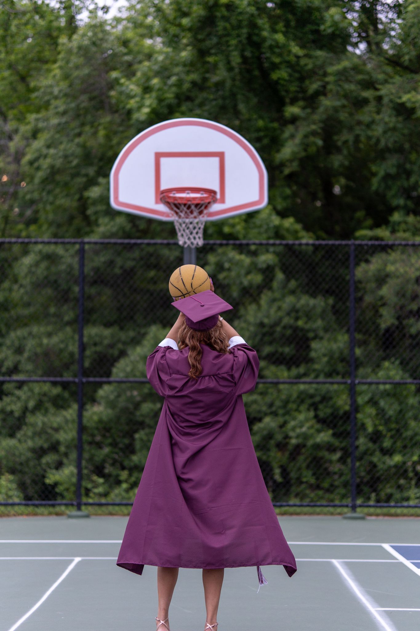 A graduate in maroon cap and gown prepares to shoot a basketball toward an outdoor hoop from behind. In Havre de grace