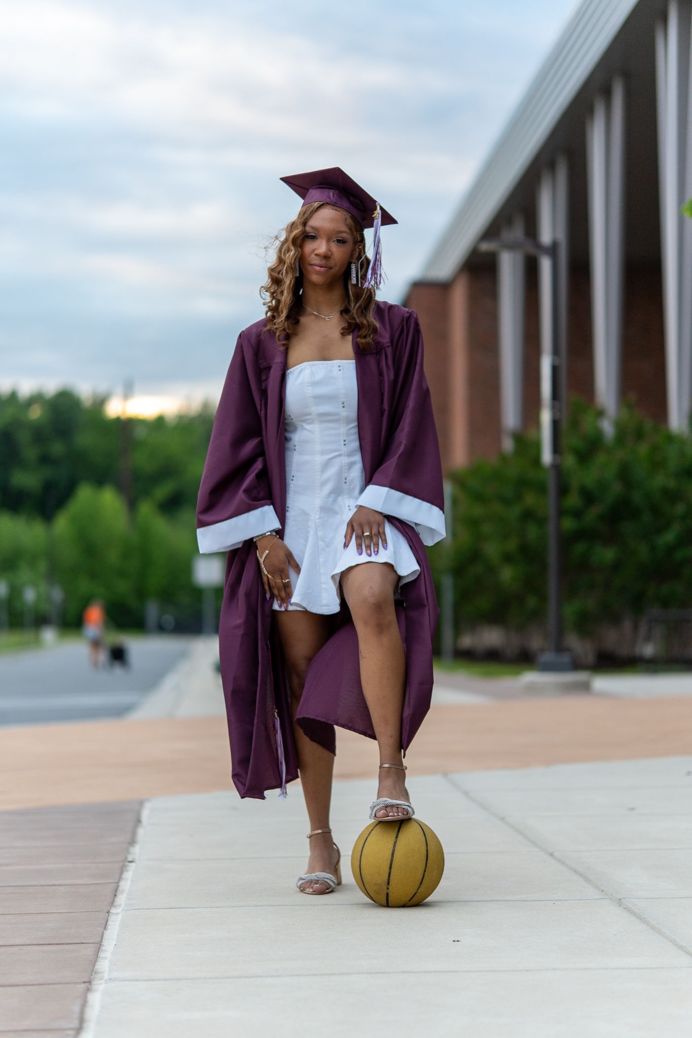 A graduating student in maroon cap and gown poses with a basketball on a campus walkway at dusk.
In Havre de Grace