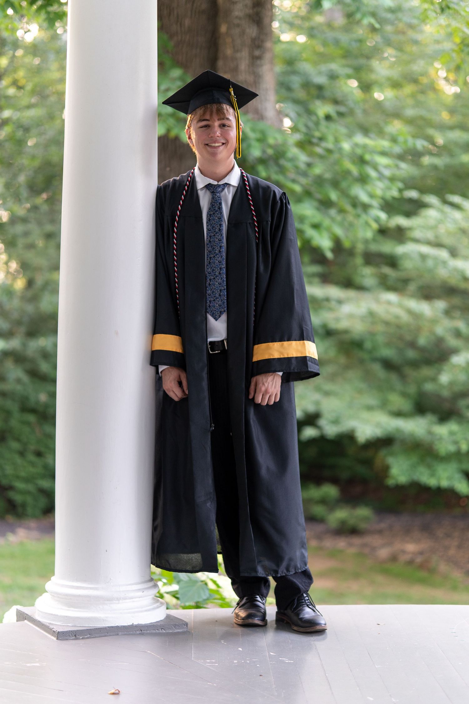 Smiling male graduate in black cap and gown leaning against a white column in an outdoor setting. In Bel Air Maryland