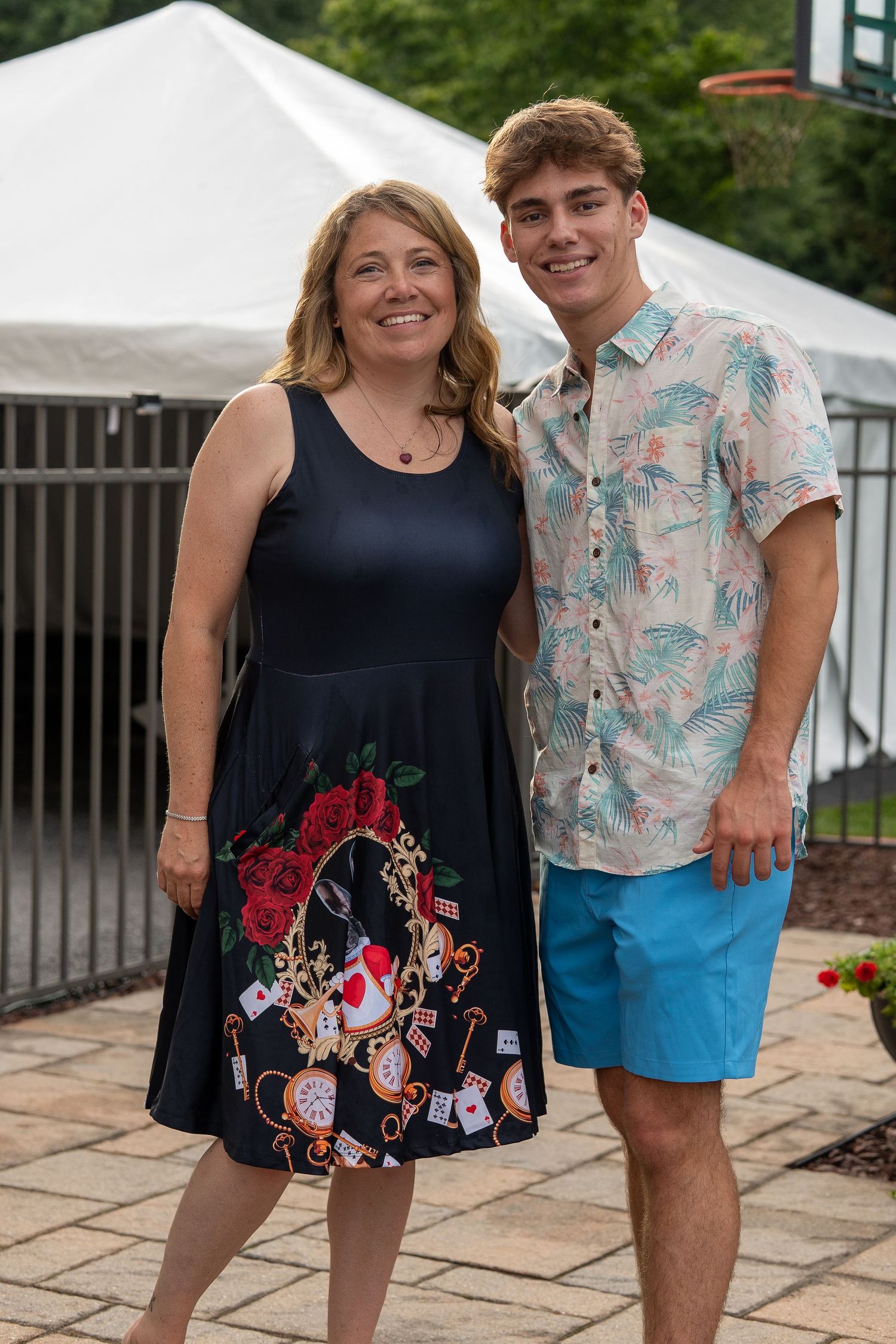 Smiling woman in black floral dress poses with tall young man in floral shirt outdoors. In Forest Hill Maryland