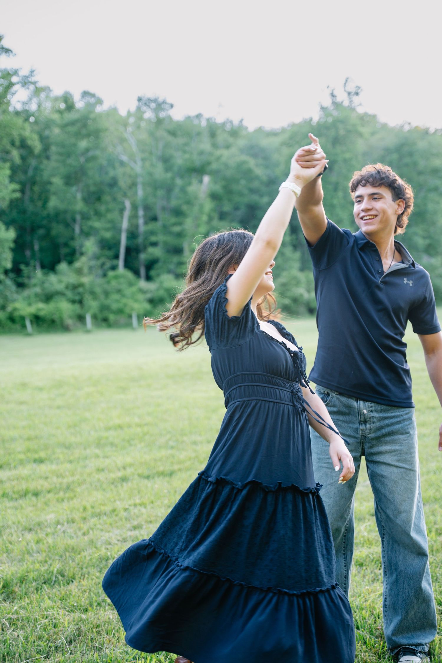 A young couple dancing outdoors on green grass, woman in navy dress being twirled by smiling man in black polo.