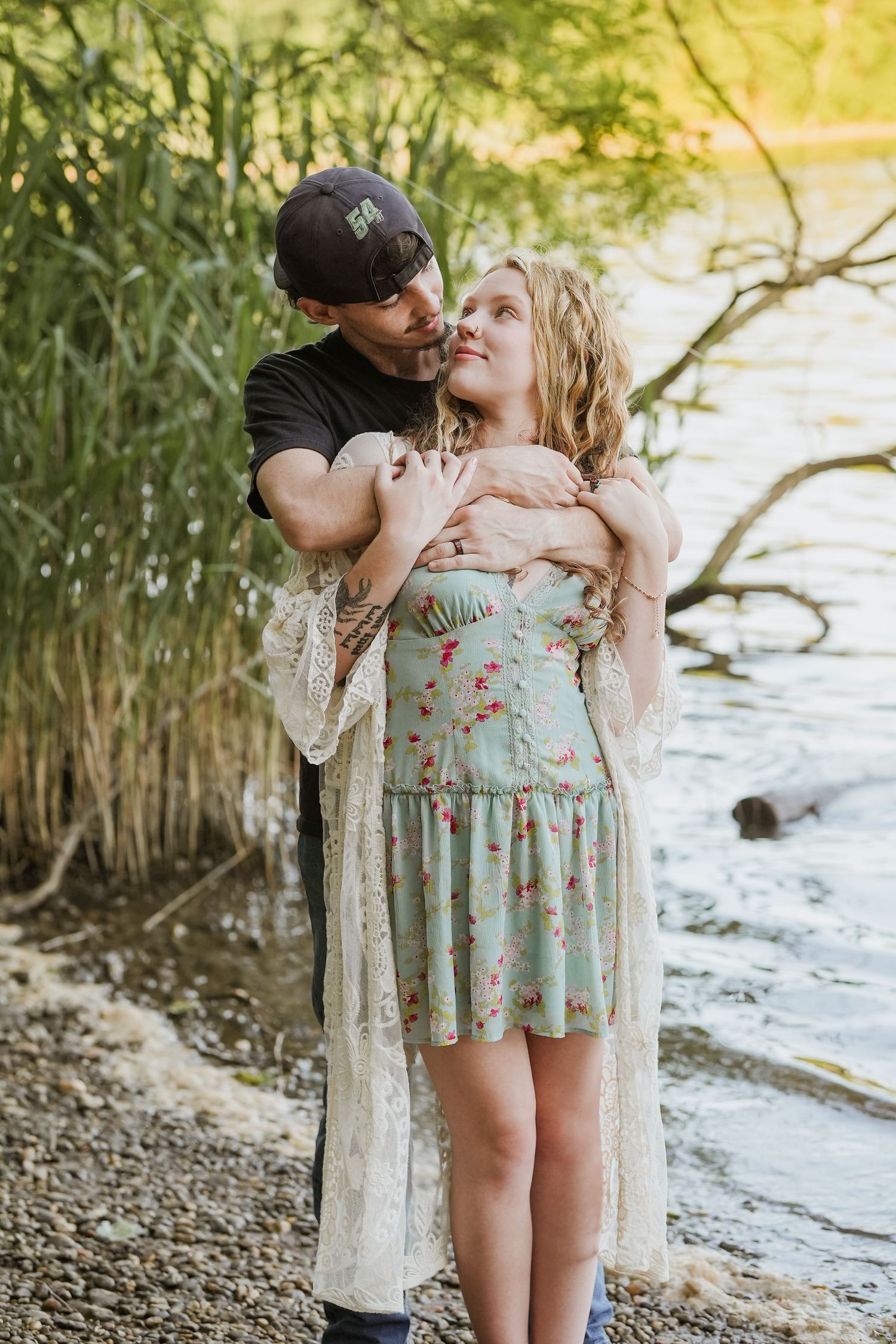 Couple embracing by a river, woman in floral dress with lace cardigan, man in black shirt and cap. In Joppatowne Maryland