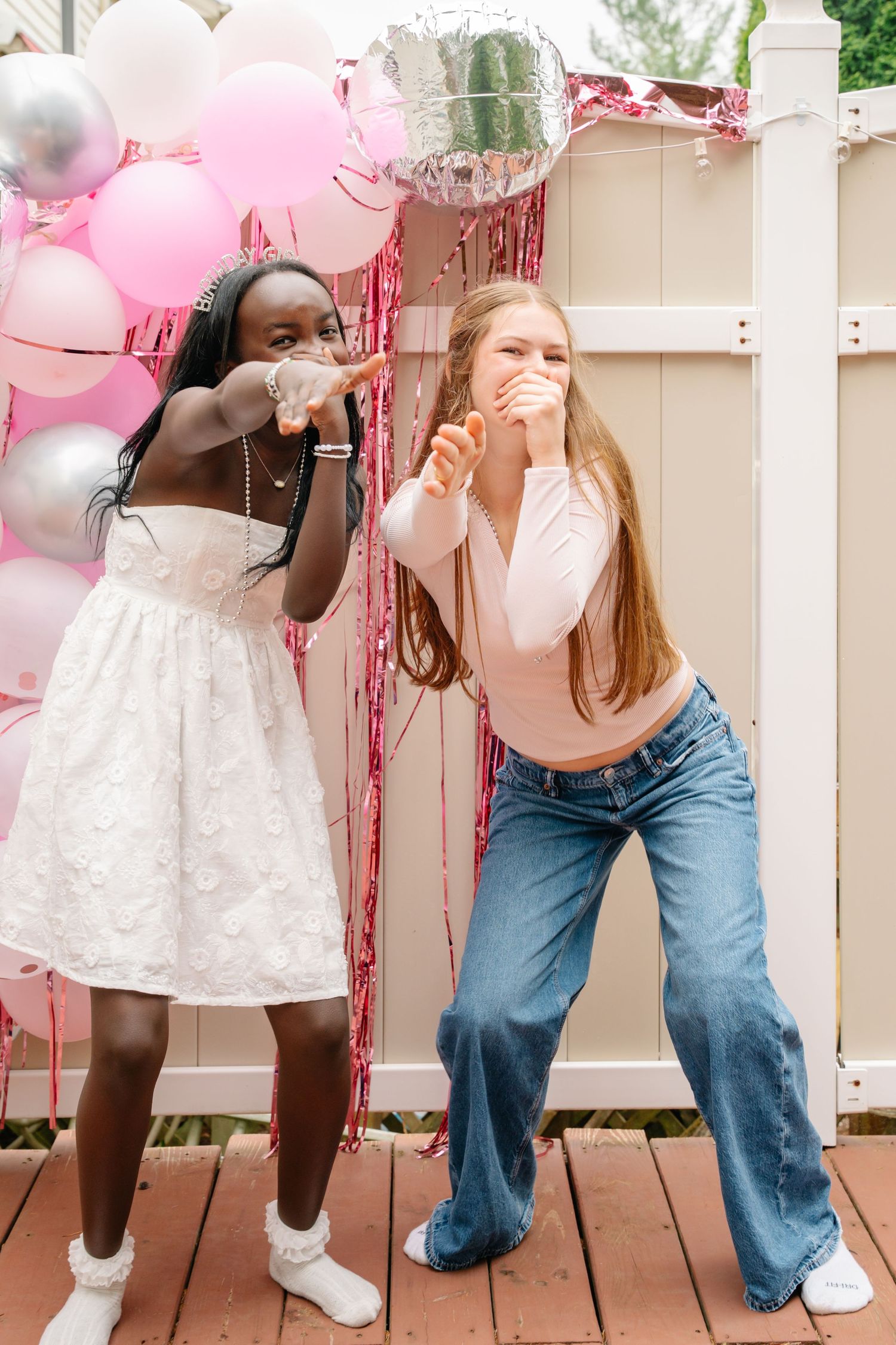 Bel Air Maryland Two girls celebrating at a pink balloon party backdrop, one in white dress, one in jeans, both laughing outdoors.