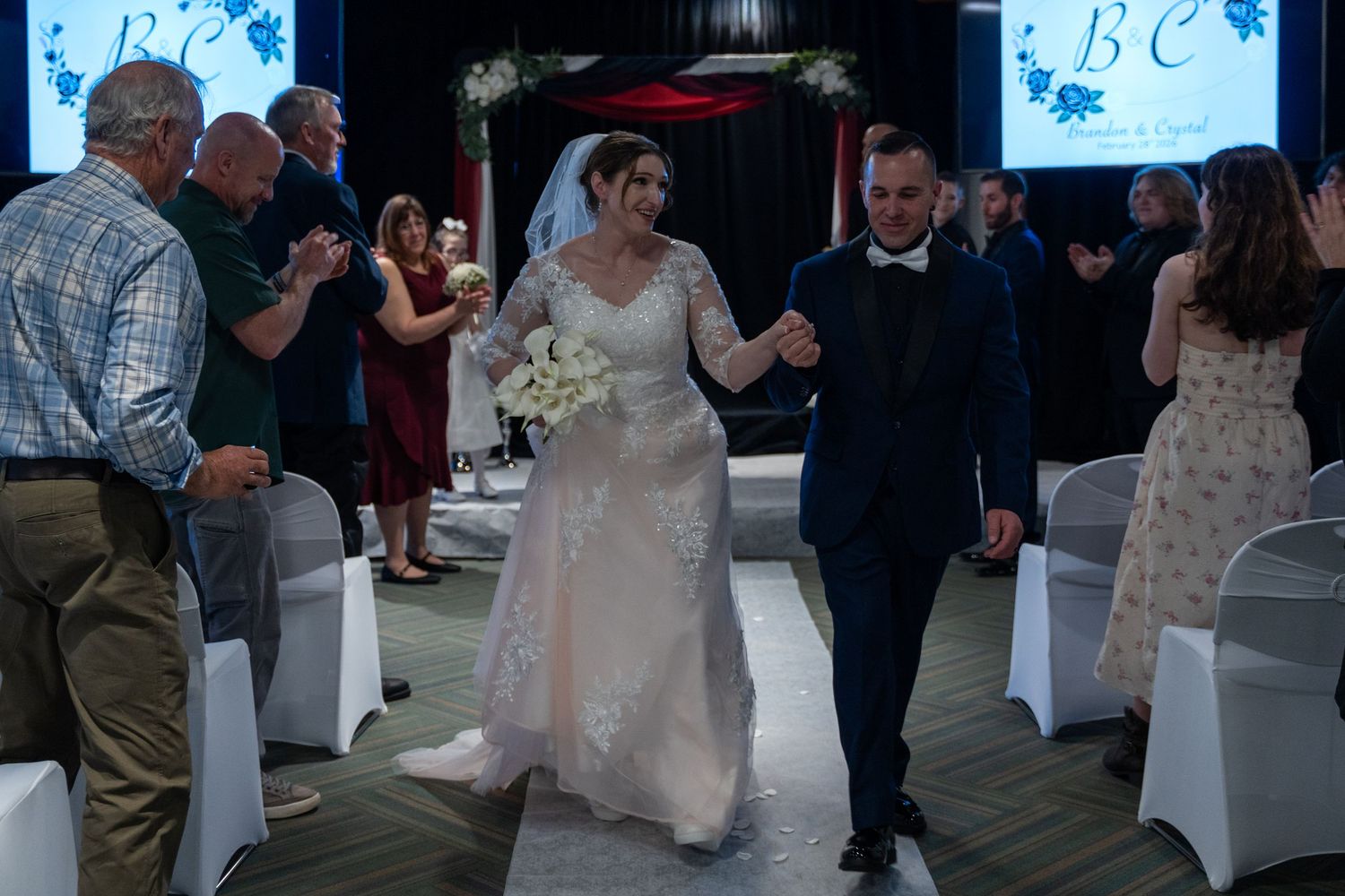 Newlyweds walk down the aisle as guests applaud at indoor wedding ceremony with floral arch decor.in Abingdon Maryland