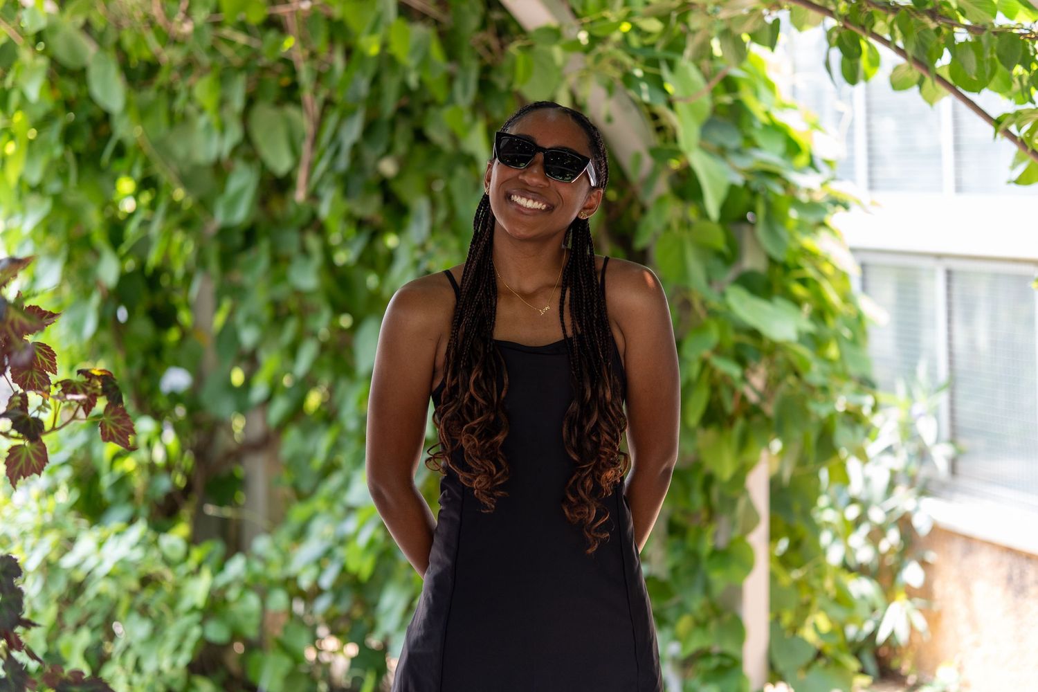Smiling woman with long braids and sunglasses in a black dress standing outdoors among green leaves. In Washington DC