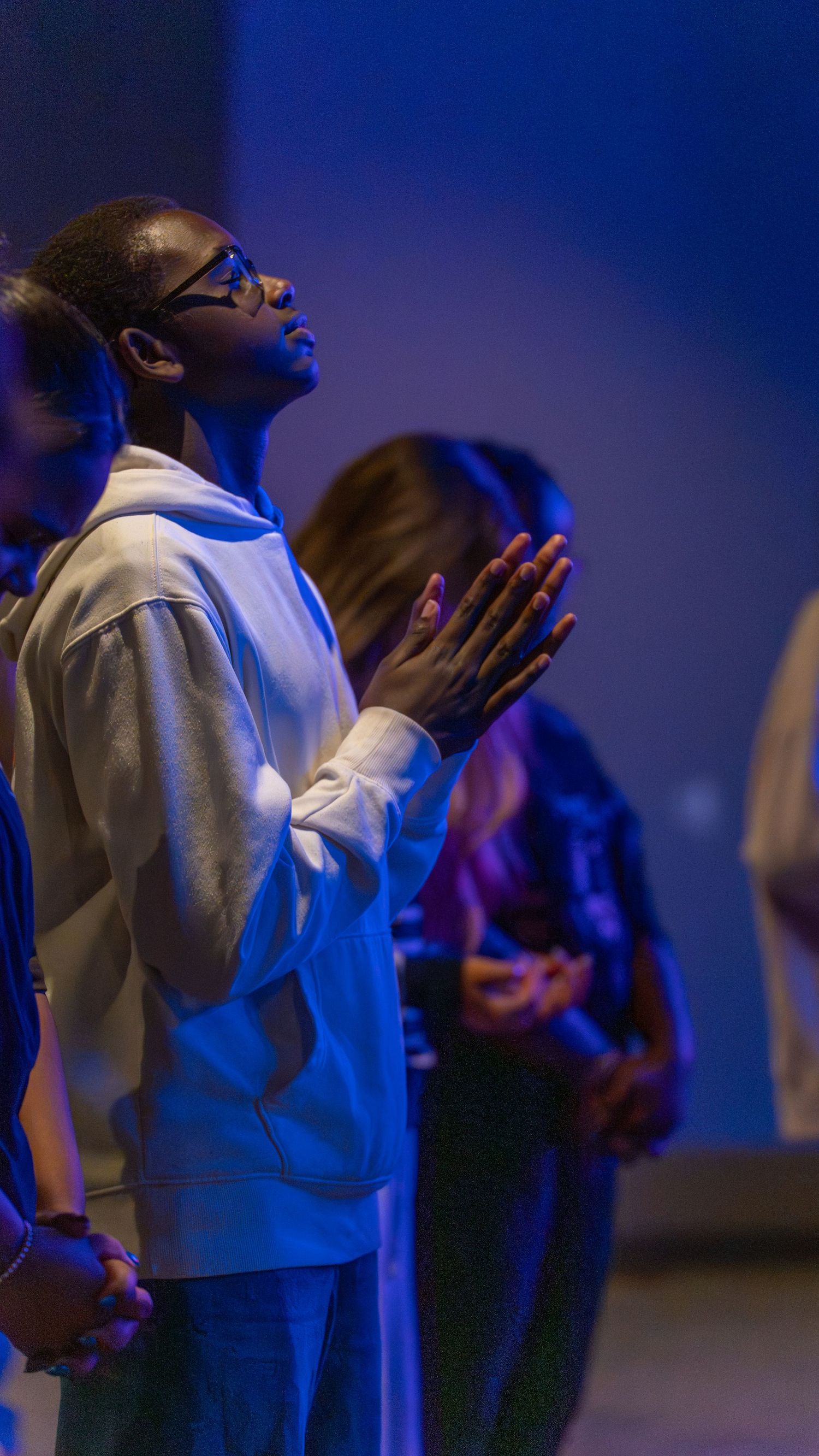 Young man in white hoodie praying with hands clasped, eyes raised upward in blue-lit worship setting.
In Abingdon Maryland