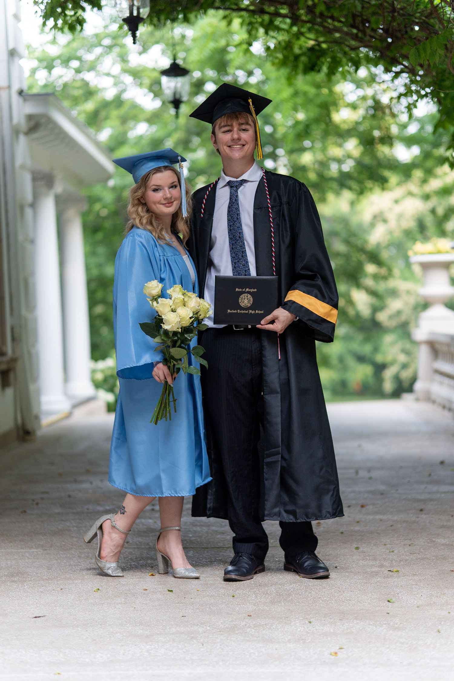 Two graduates in cap and gown posing together outdoors, woman holding white roses and man holding diploma. In Bel Air Maryland
