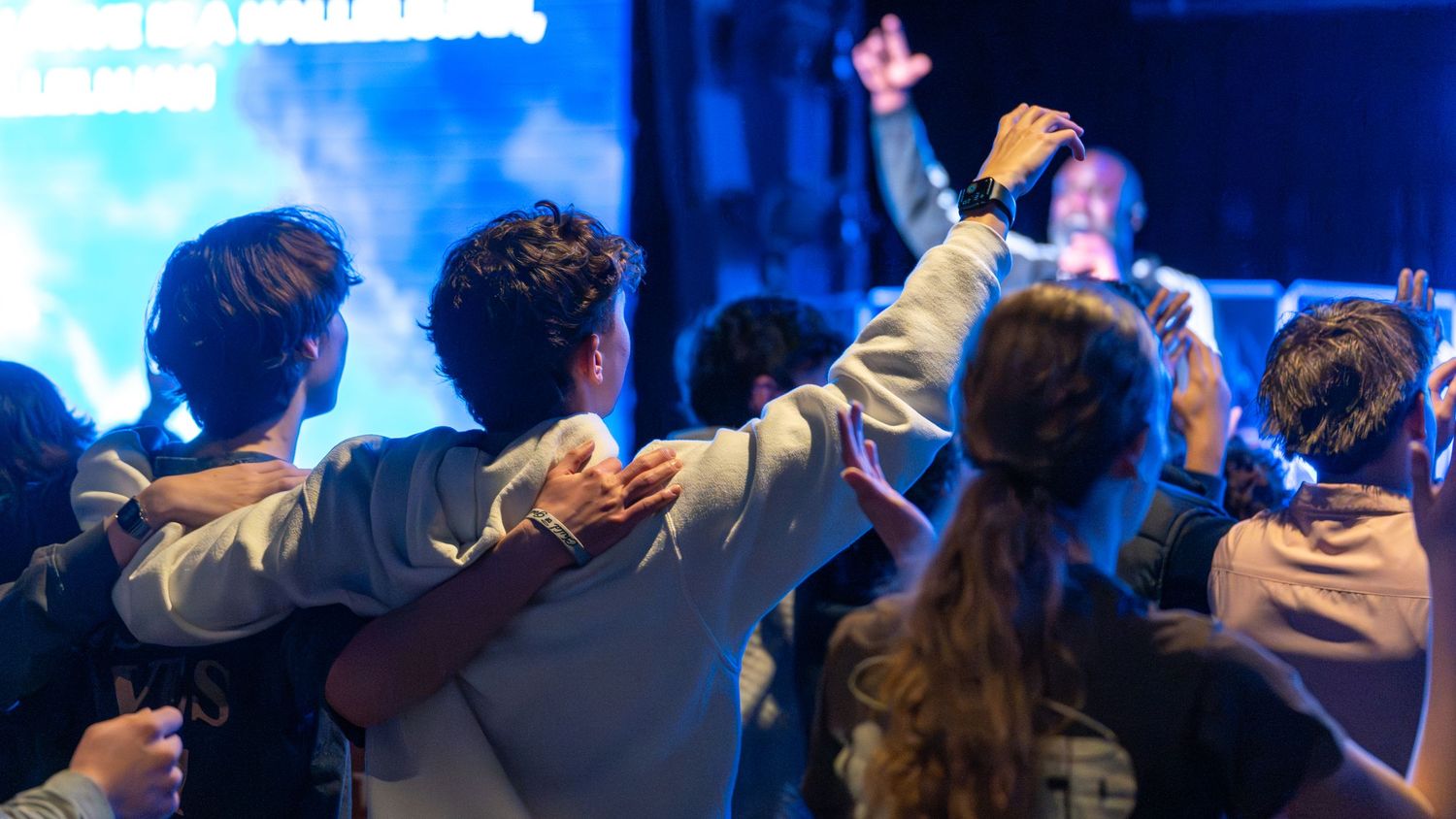 Group of young people with arms around each other facing a lit stage during a worship event. In Abingdon Maryland