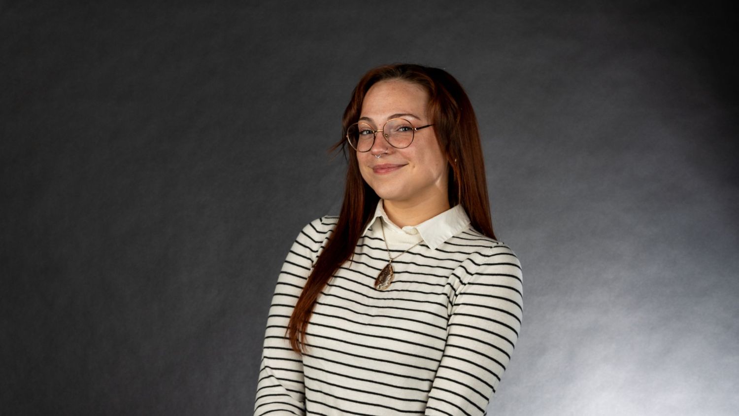 Young woman with long brown hair and glasses smiling, wearing a striped turtleneck against a gray background. In Bel Air Maryland