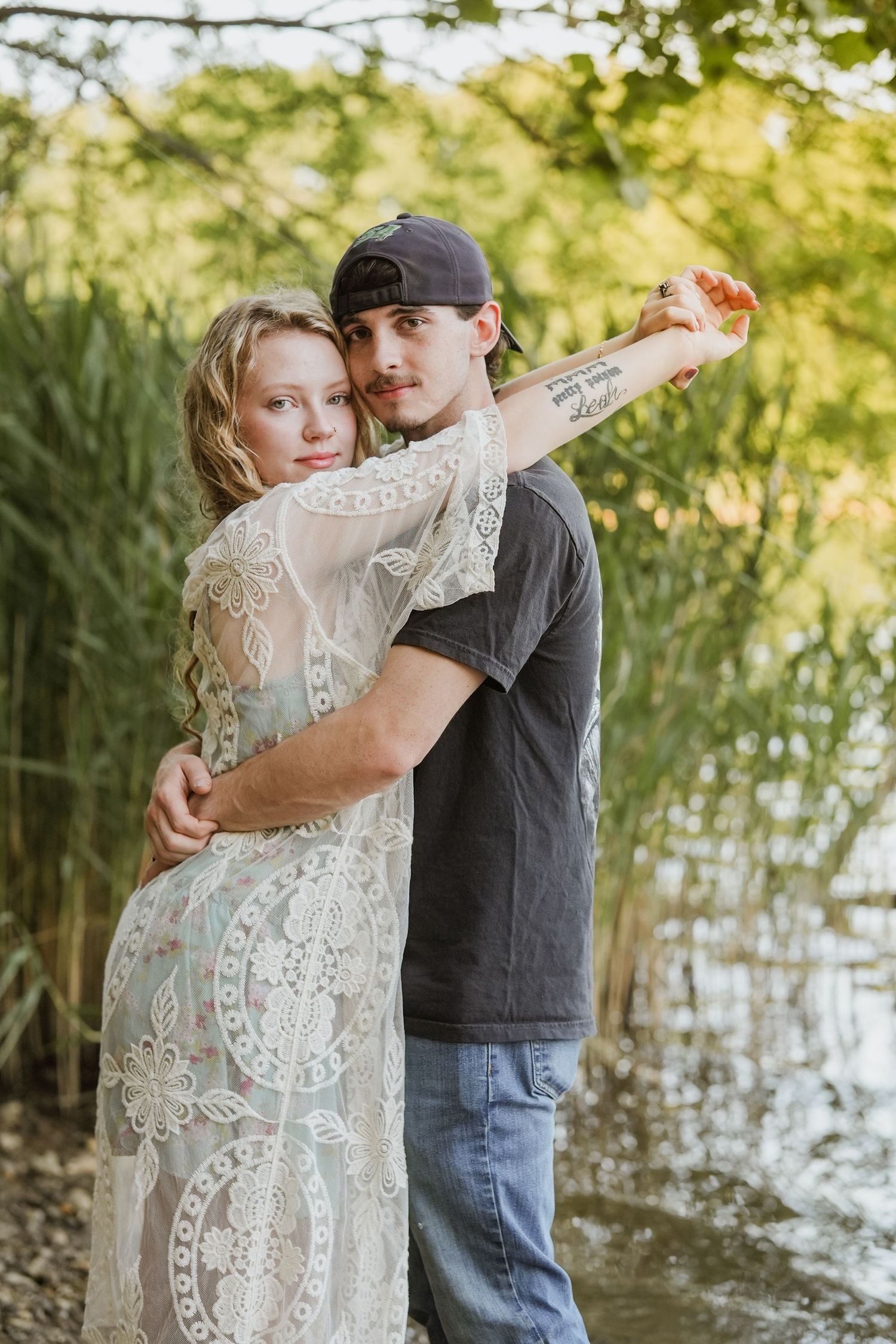 A couple embraces outdoors near a lake, woman in lace dress, man in gray tee and cap, surrounded by greenery.