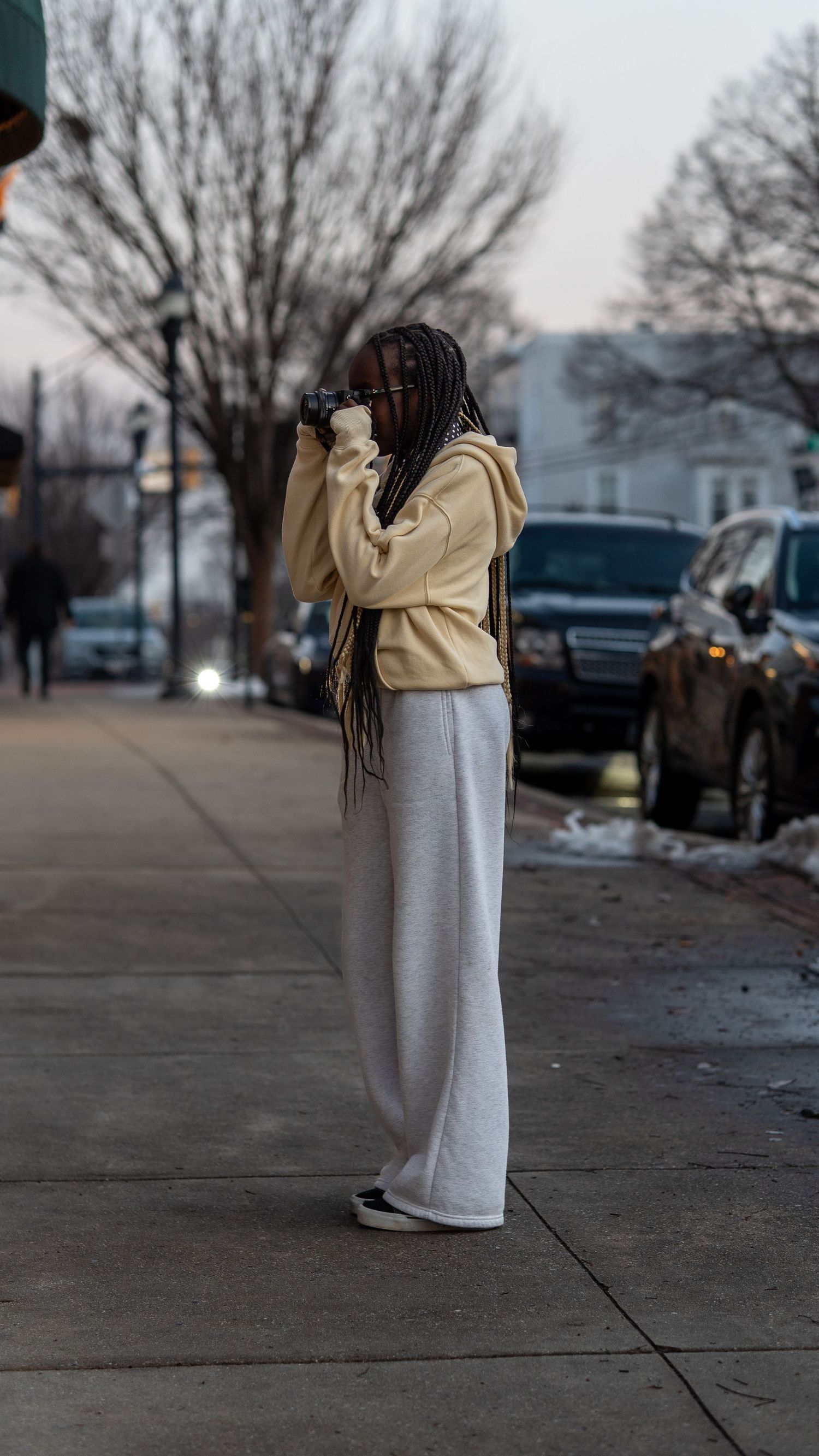 Stylish woman with long braids takes photos with camera on city sidewalk at dusk in casual outfit.In Bel Air Maryland