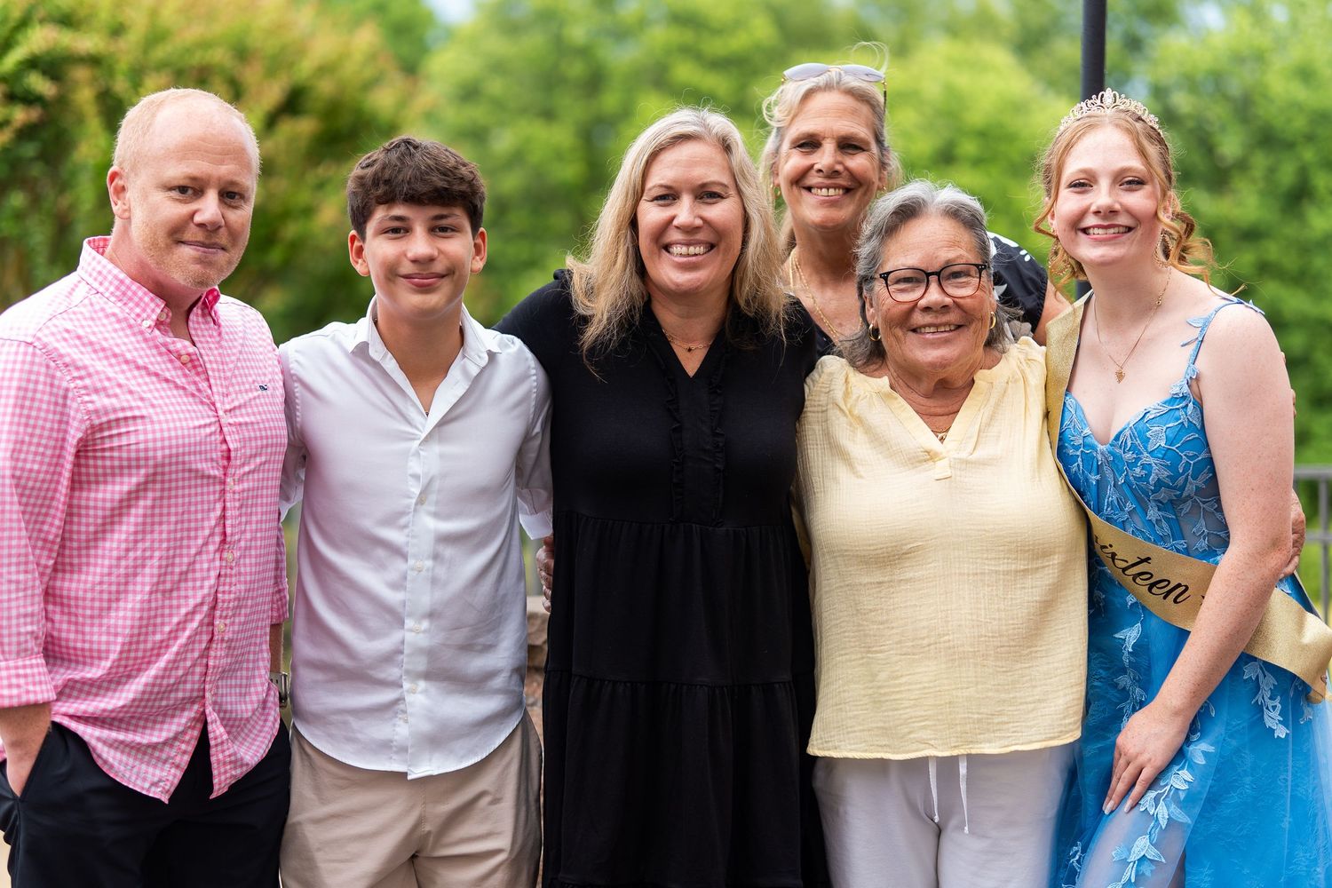 Family of six smiling outdoors, including teen girl in blue formal gown with Sweet Sixteen sash. In Forest Hill Maryland
