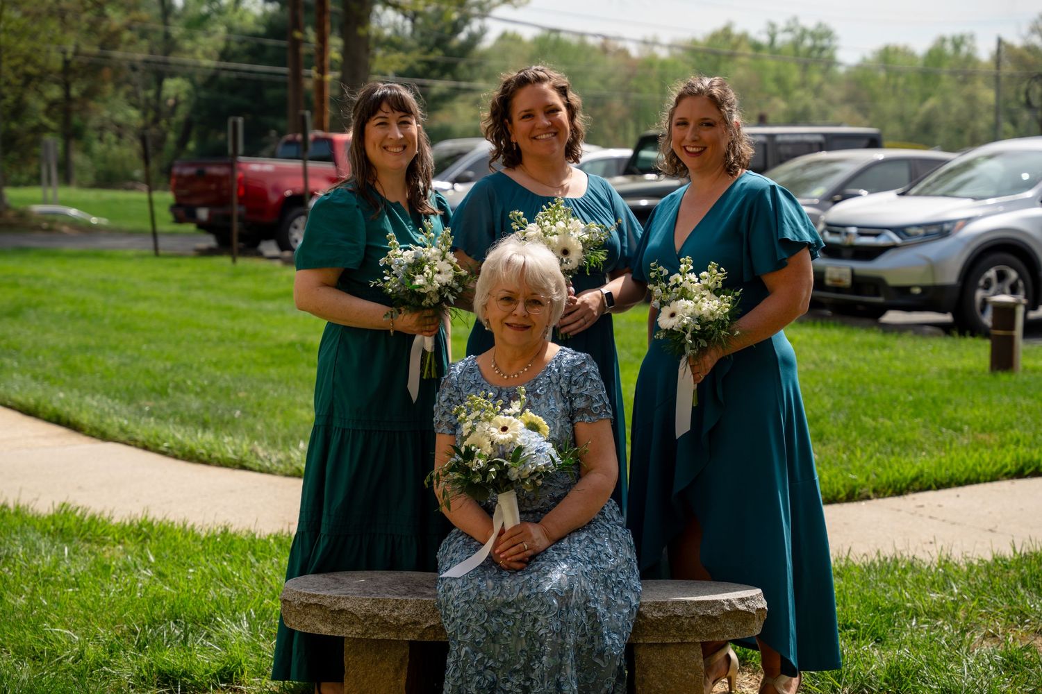 in Bel air MD Three bridesmaids in teal dresses holding white bouquets stand behind an elderly woman seated on a stone bench outdoors.