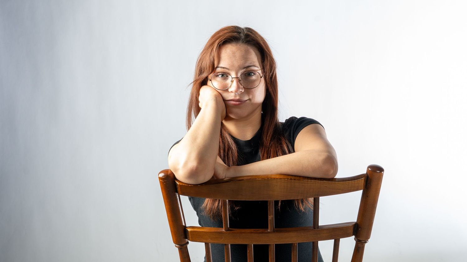 Young woman with red hair and glasses resting chin on arms over back of wooden chair, looking at camera.  In Bel Air Maryland