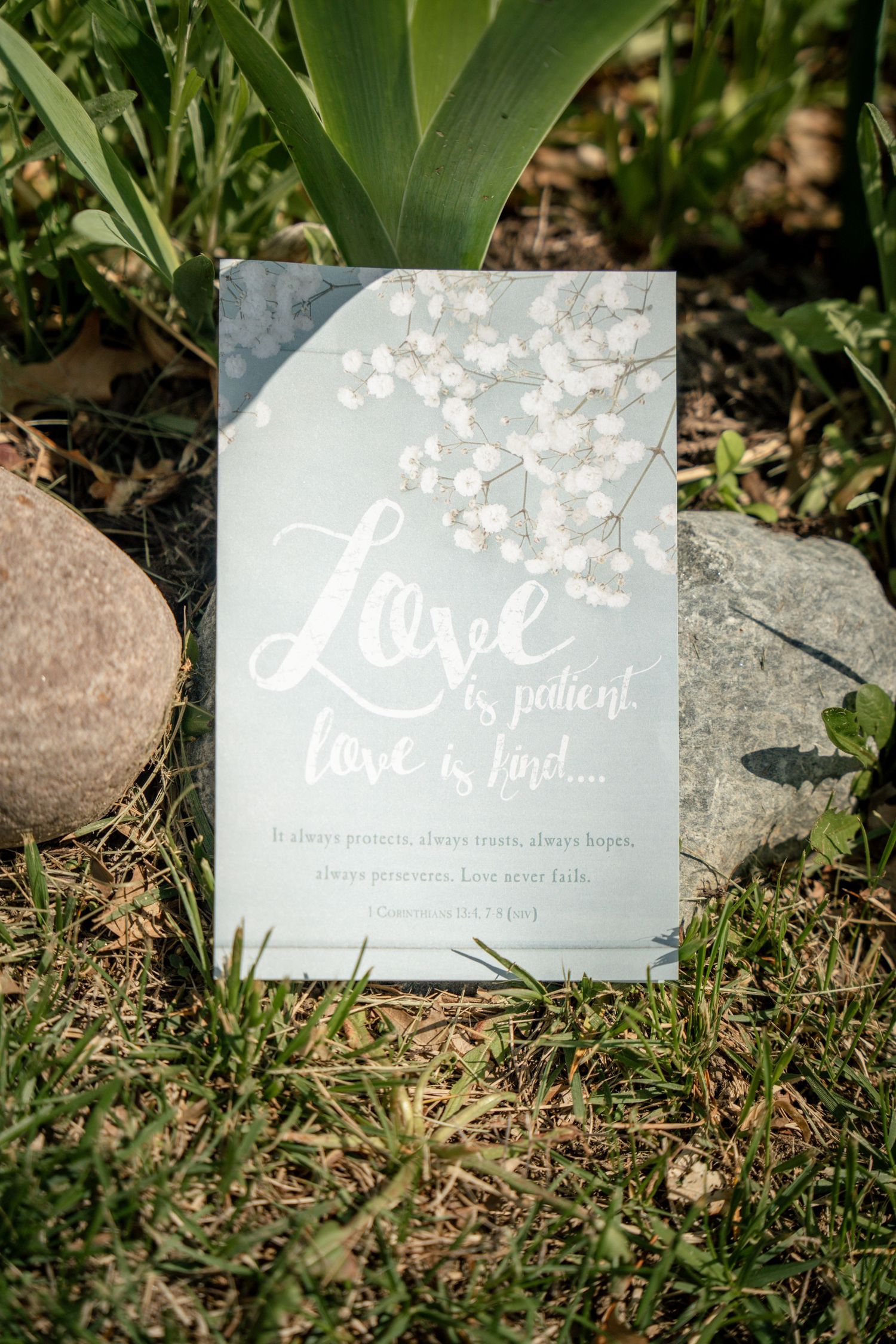 A decorative wedding sign reading Love is Patient, Love is Kind with floral design, placed among rocks and greenery. In Bel Air Maryland