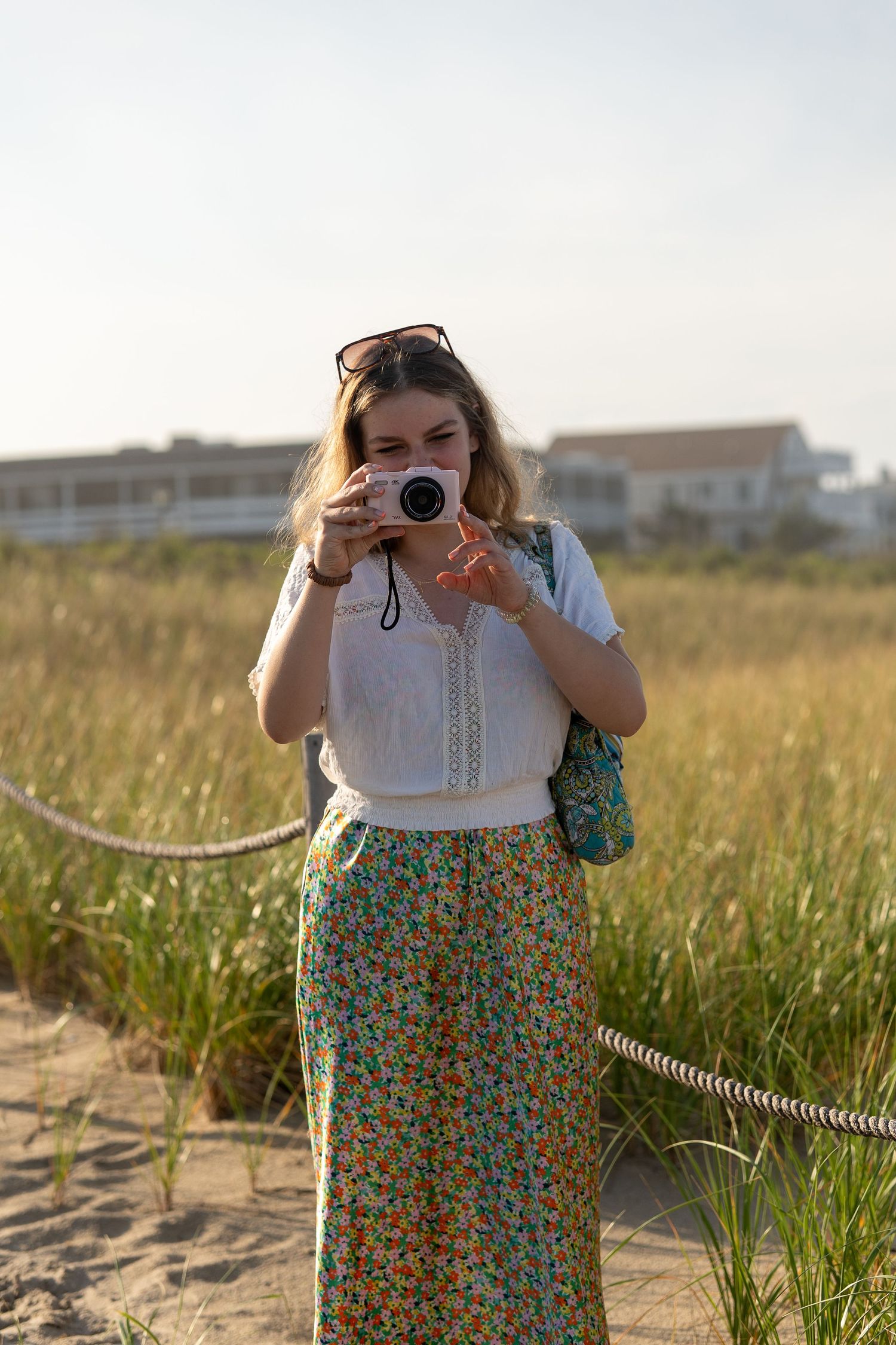 A young woman in a floral skirt photographs with a white camera on a sandy coastal path surrounded by tall grass. At Bethany beach Delaware