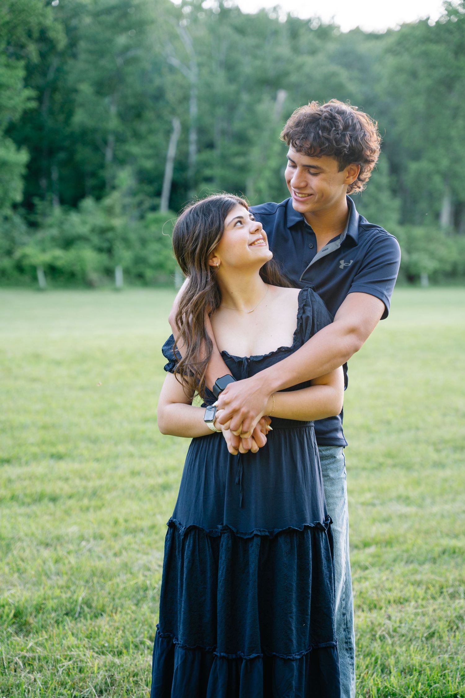 A young couple embraces in a green field, the woman in a black dress smiling up at the man holding her hands.