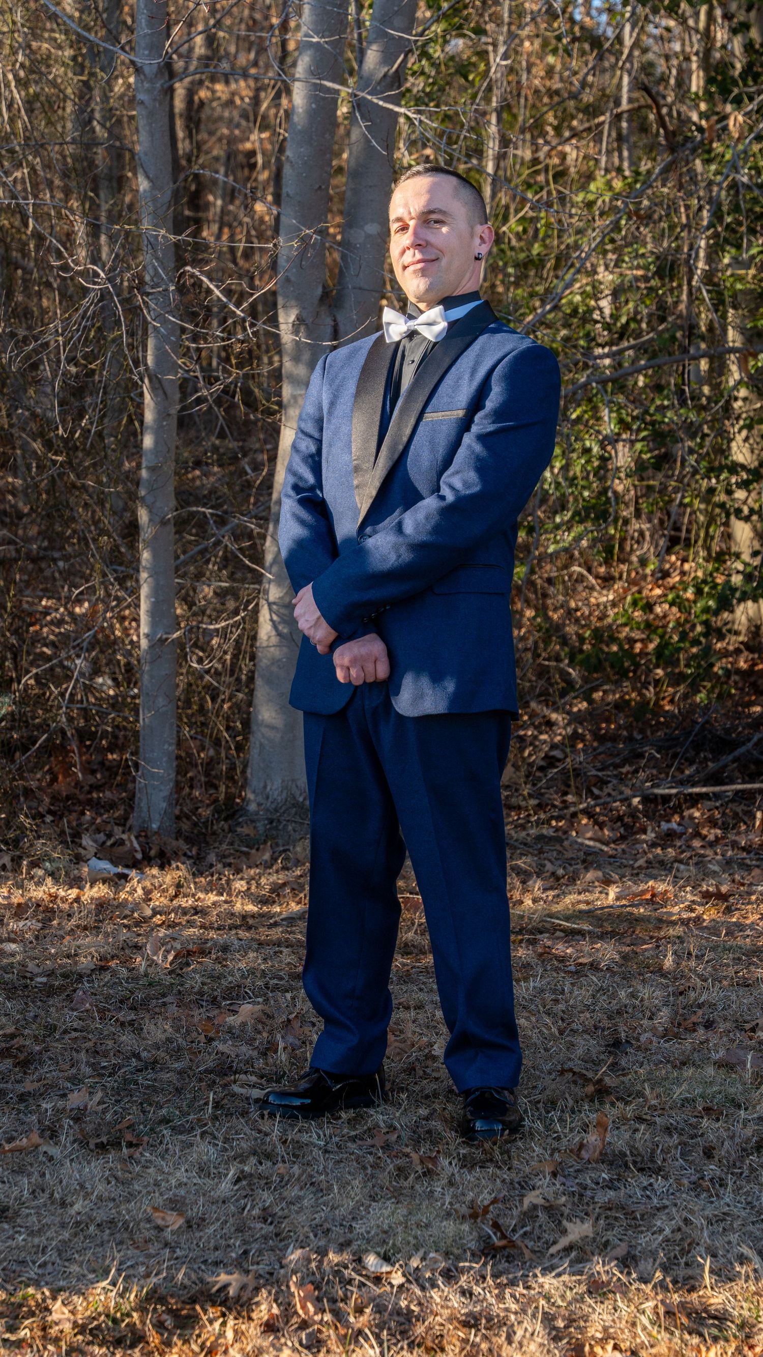 Groom in navy blue tuxedo with gold bow tie stands confidently in a sunlit autumn forest setting.in Abingdon Maryland