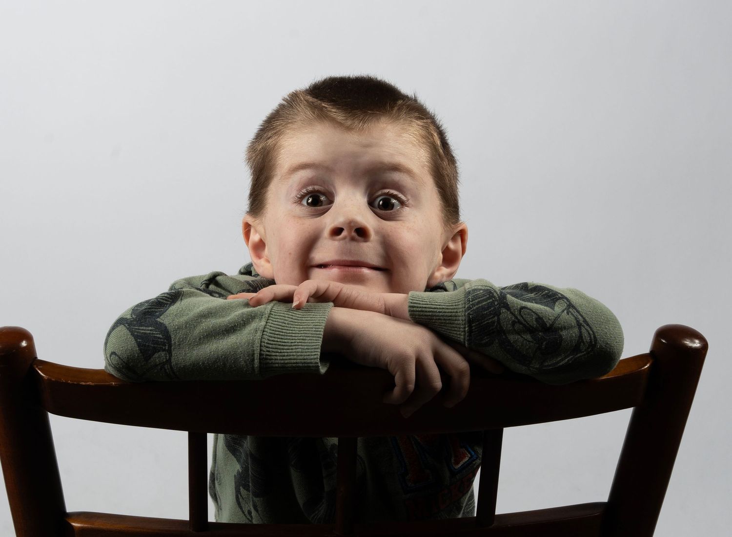 A cheerful young boy rests his chin on his arms over the back of a wooden chair, smiling widely. In Bel Air Maryland