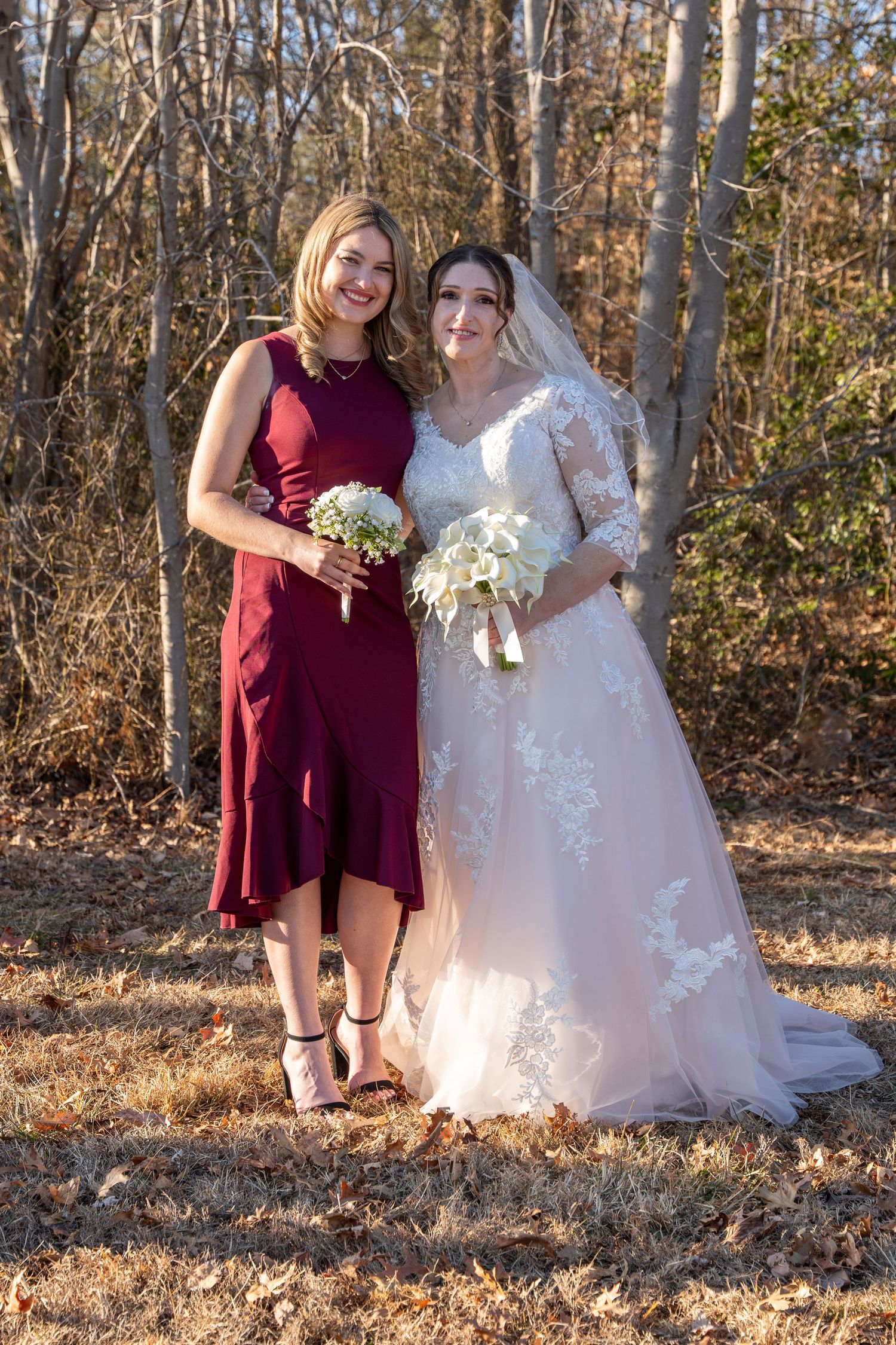 Bride in lace wedding gown poses with bridesmaid in burgundy dress holding bouquets outdoors in autumn forest.in Abingdon Maryland