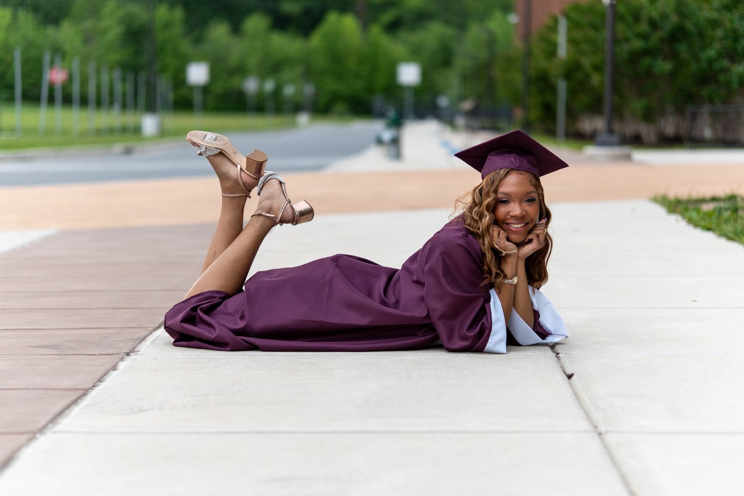 A graduate in a maroon cap and gown lies on a sidewalk smiling during an outdoor graduation photo session. In Havre de grace