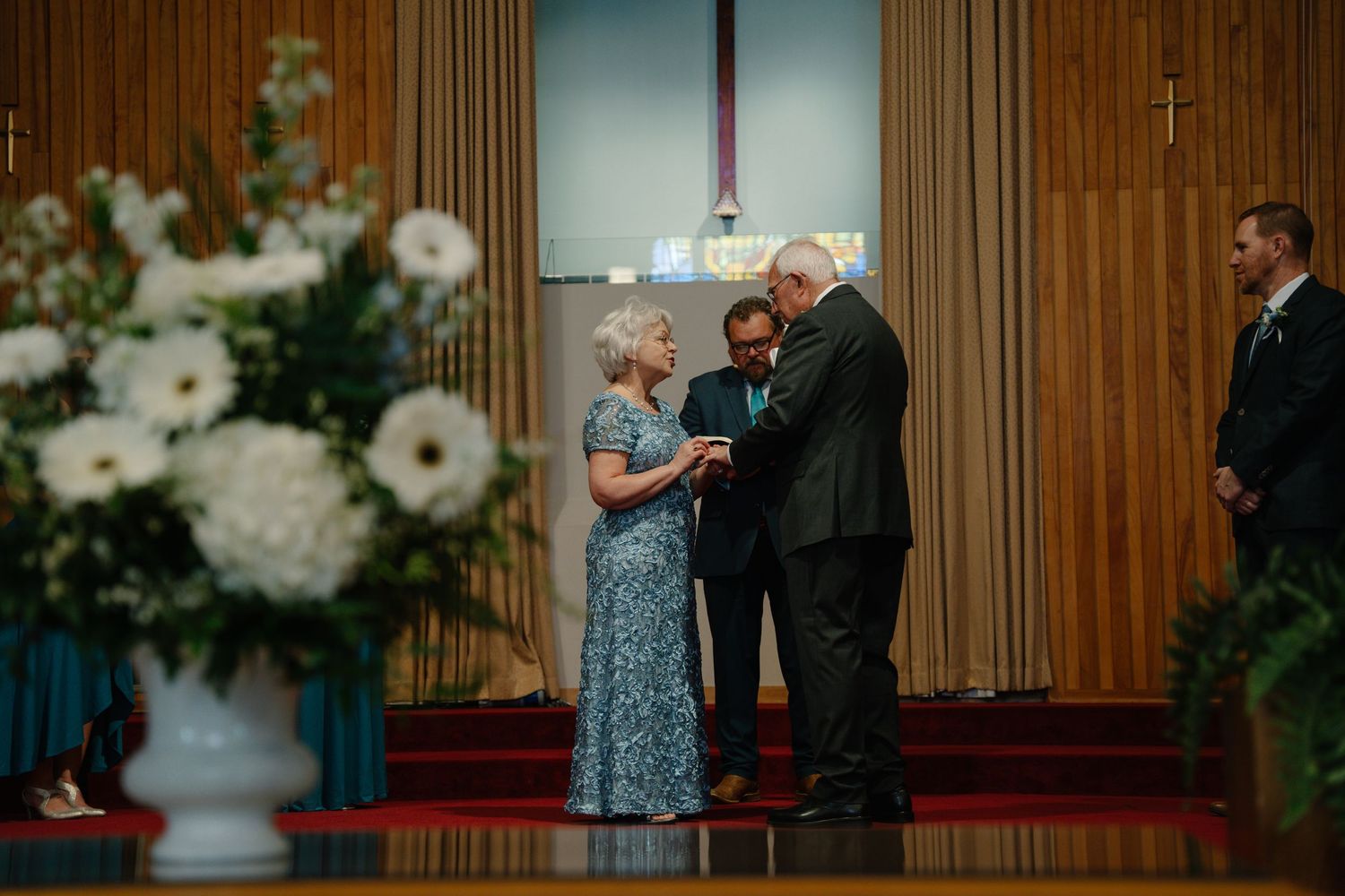 Elderly couple exchanging vows at a church wedding ceremony with white floral arrangements and officiant. In Bel air Maryland