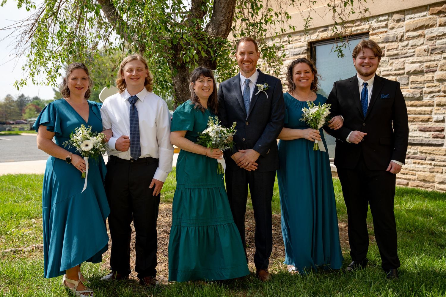 In Bel Air Maryland Wedding party in teal dresses and suits posing outdoors, smiling with white floral bouquets near a stone building.