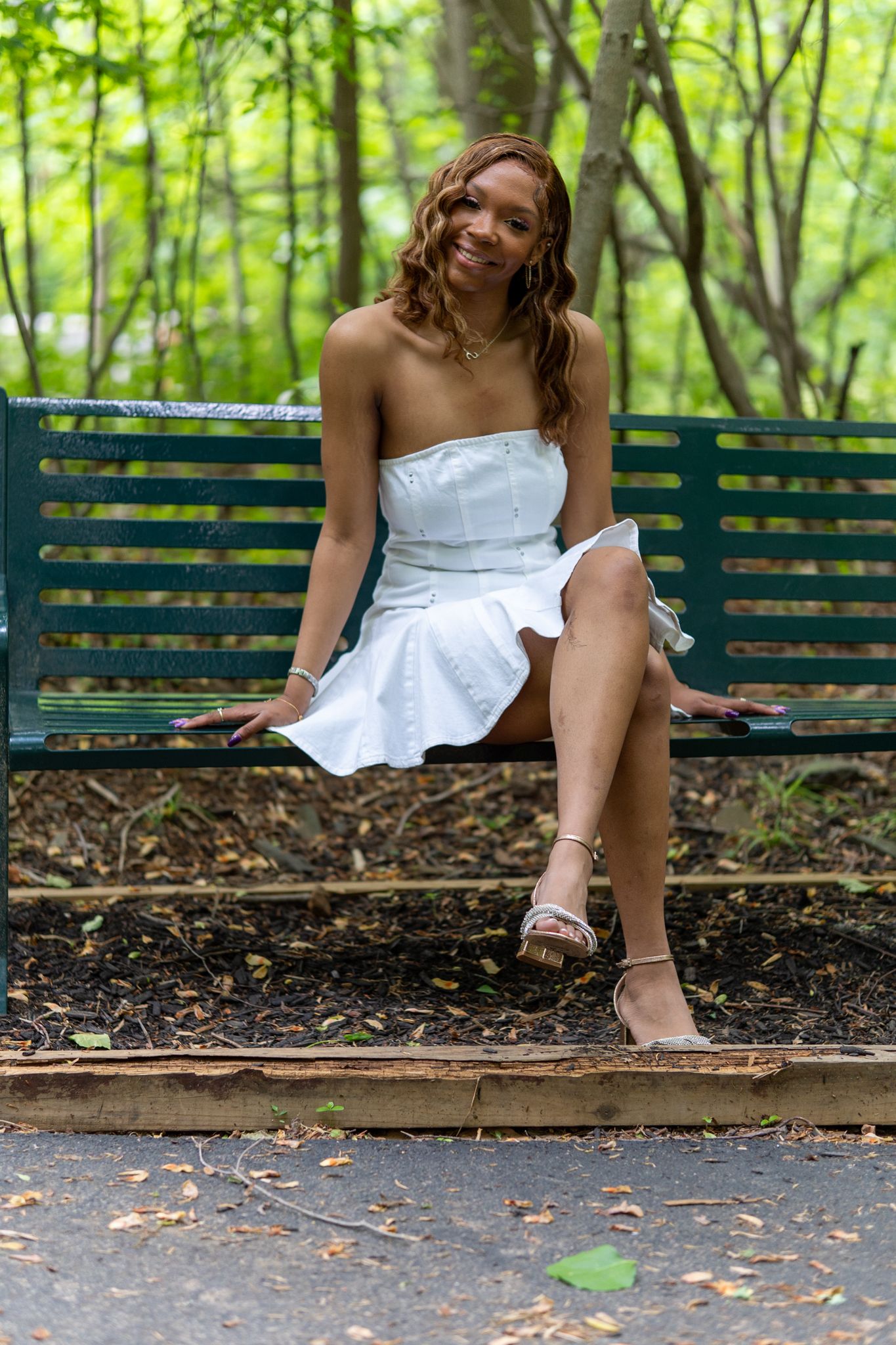 A smiling young woman in a white strapless mini dress sits on a green park bench surrounded by lush green trees. In Havre de grace Maryland
