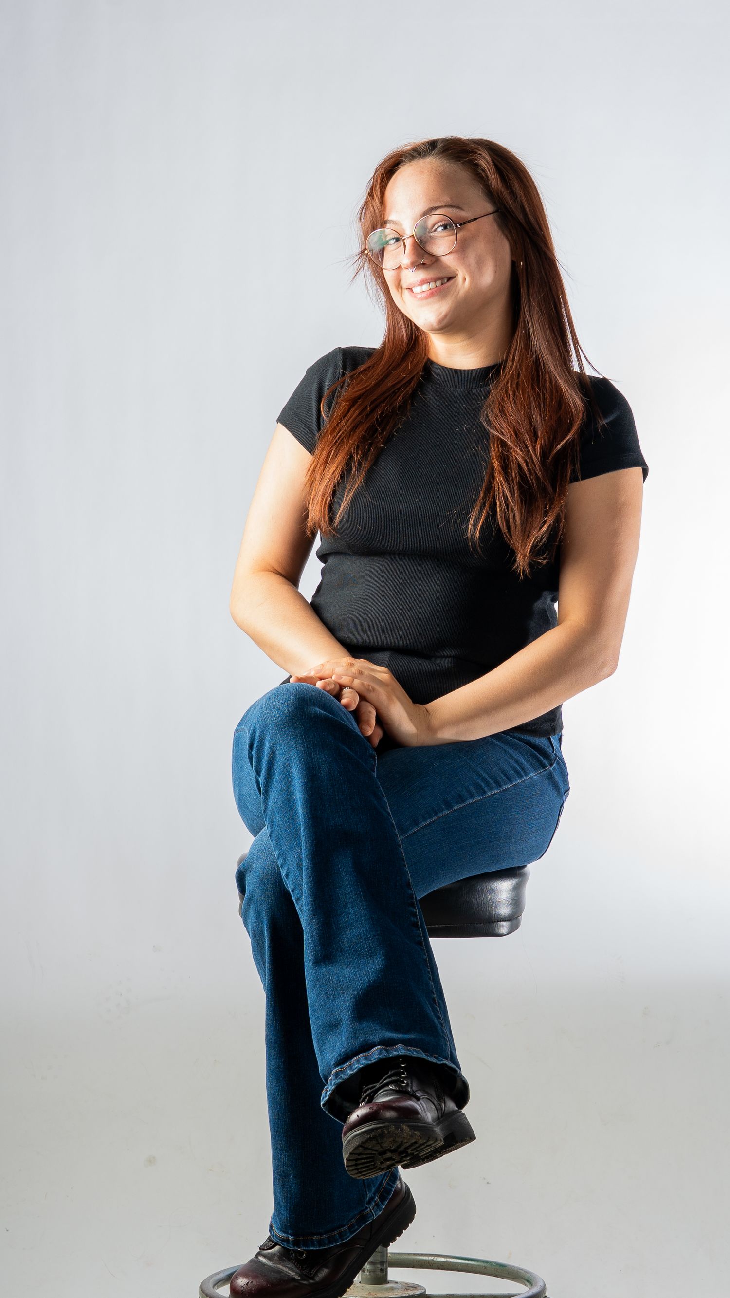 A young woman with long red hair and glasses sits on a stool smiling against a white studio background. In Bel Air