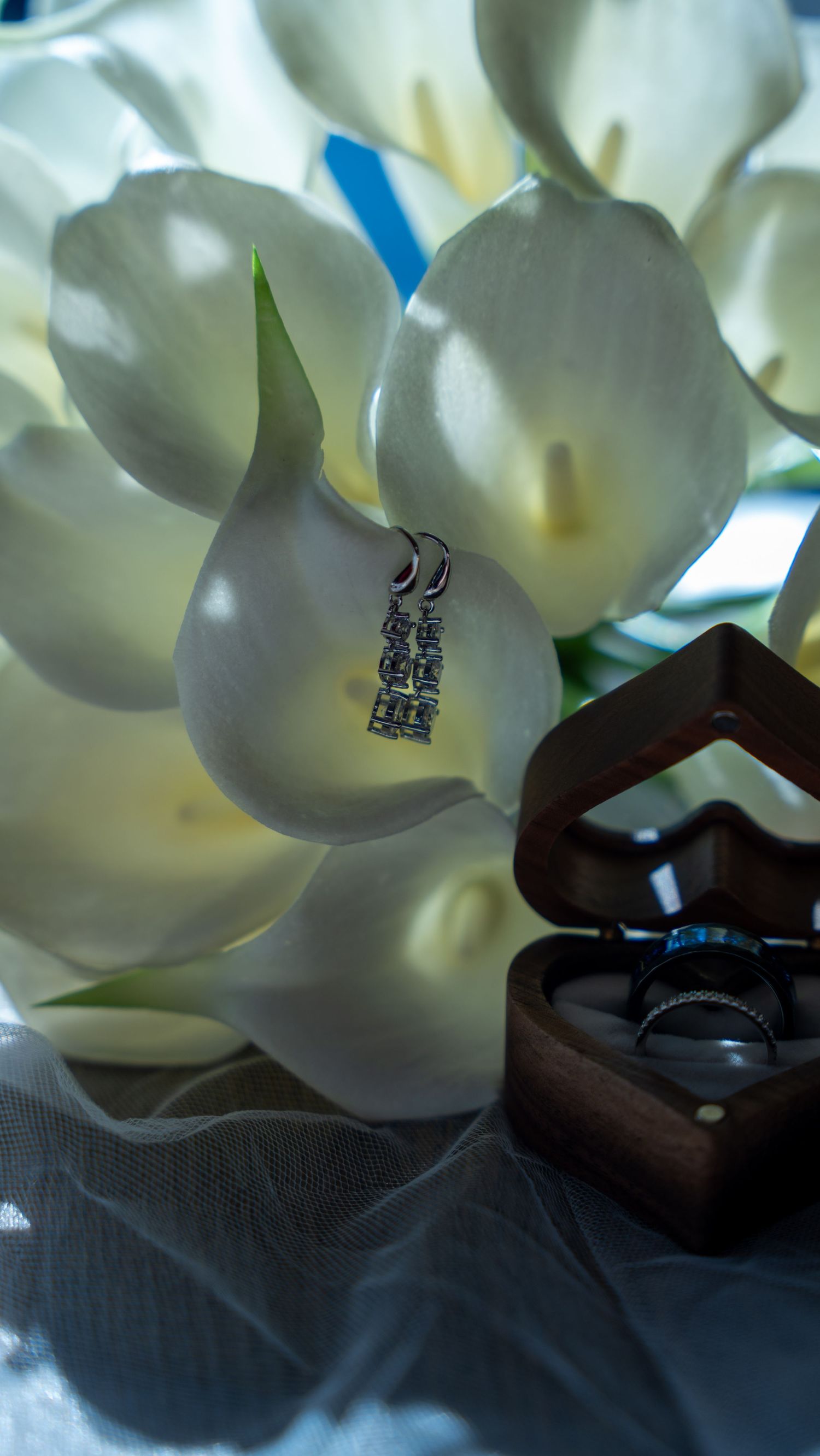 Wedding rings in a wooden heart box beside white calla lilies and a diamond pendant. In Abingdon Maryland