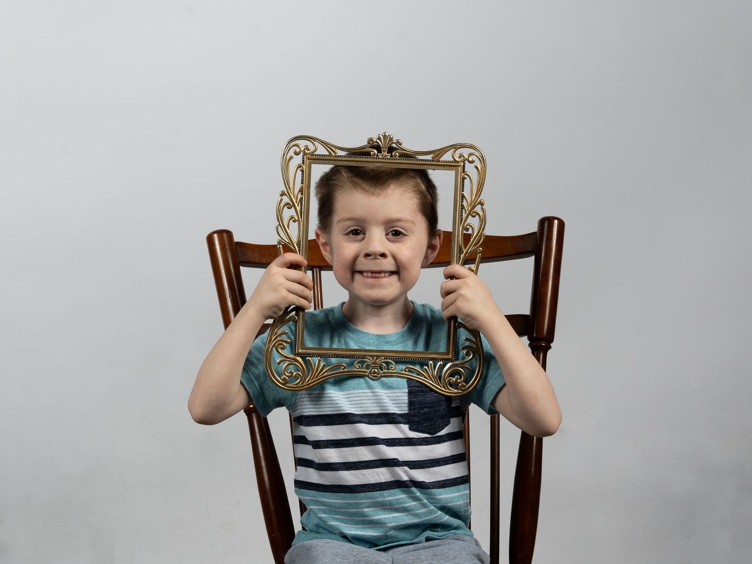 A smiling young boy holds a decorative golden picture frame around his face while sitting in a wooden chair. In Bel Air Maryland