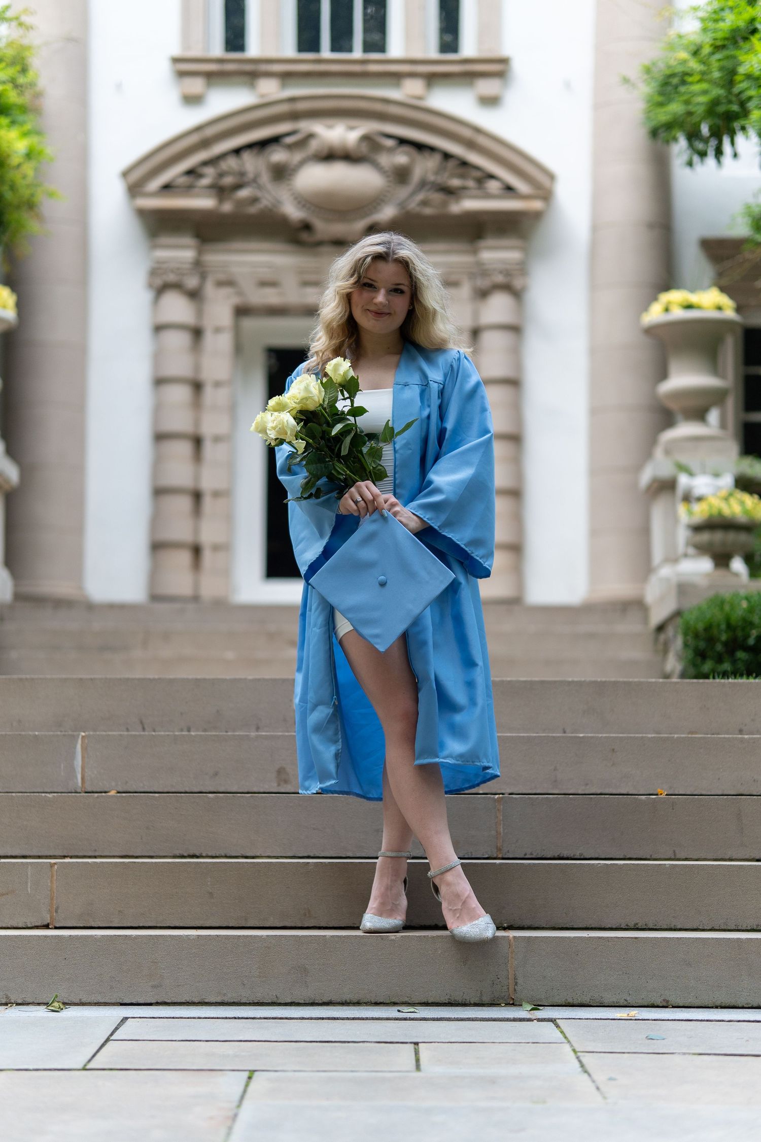 Female graduate in light blue cap and gown holding white roses on stone steps of a classical building. In Bel Air Maryland