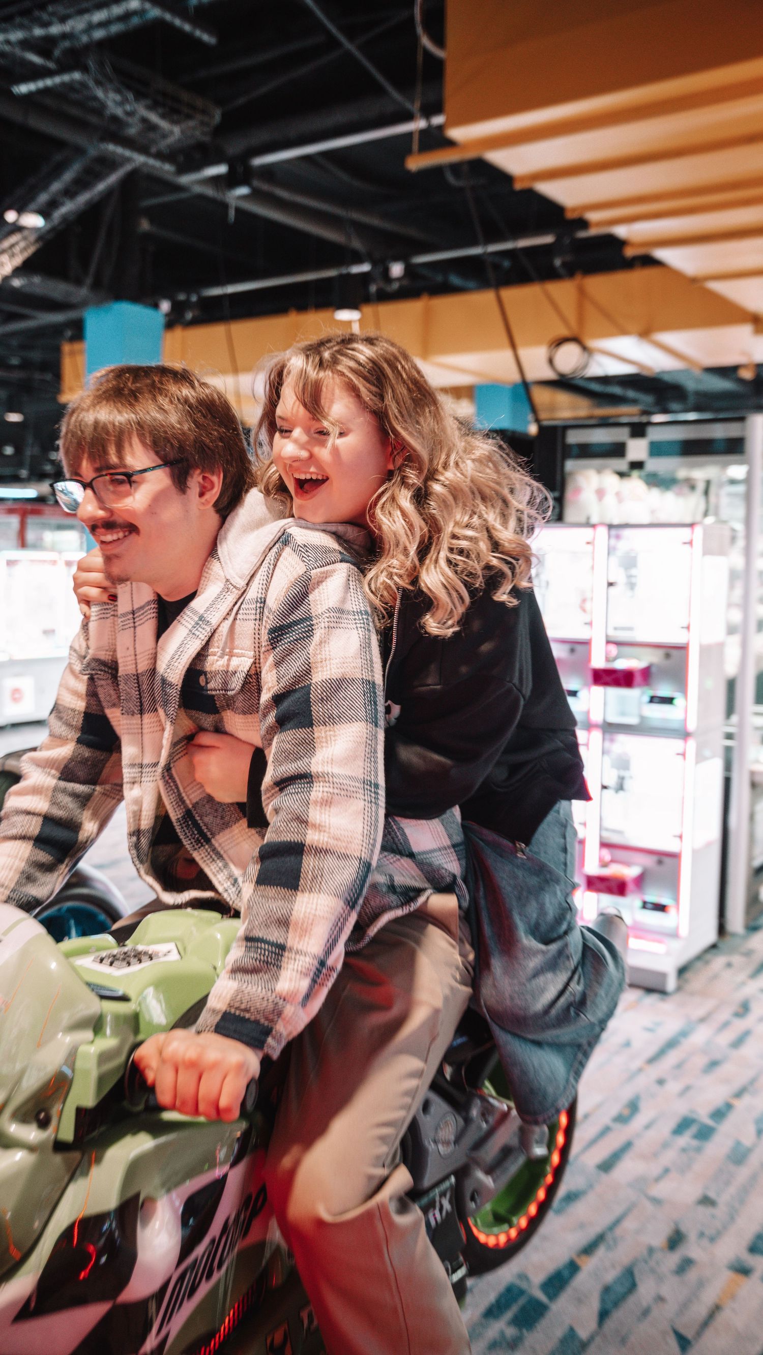 Two friends laugh and pose on an arcade motorcycle ride inside a colorful entertainment venue. In Towson Maryland