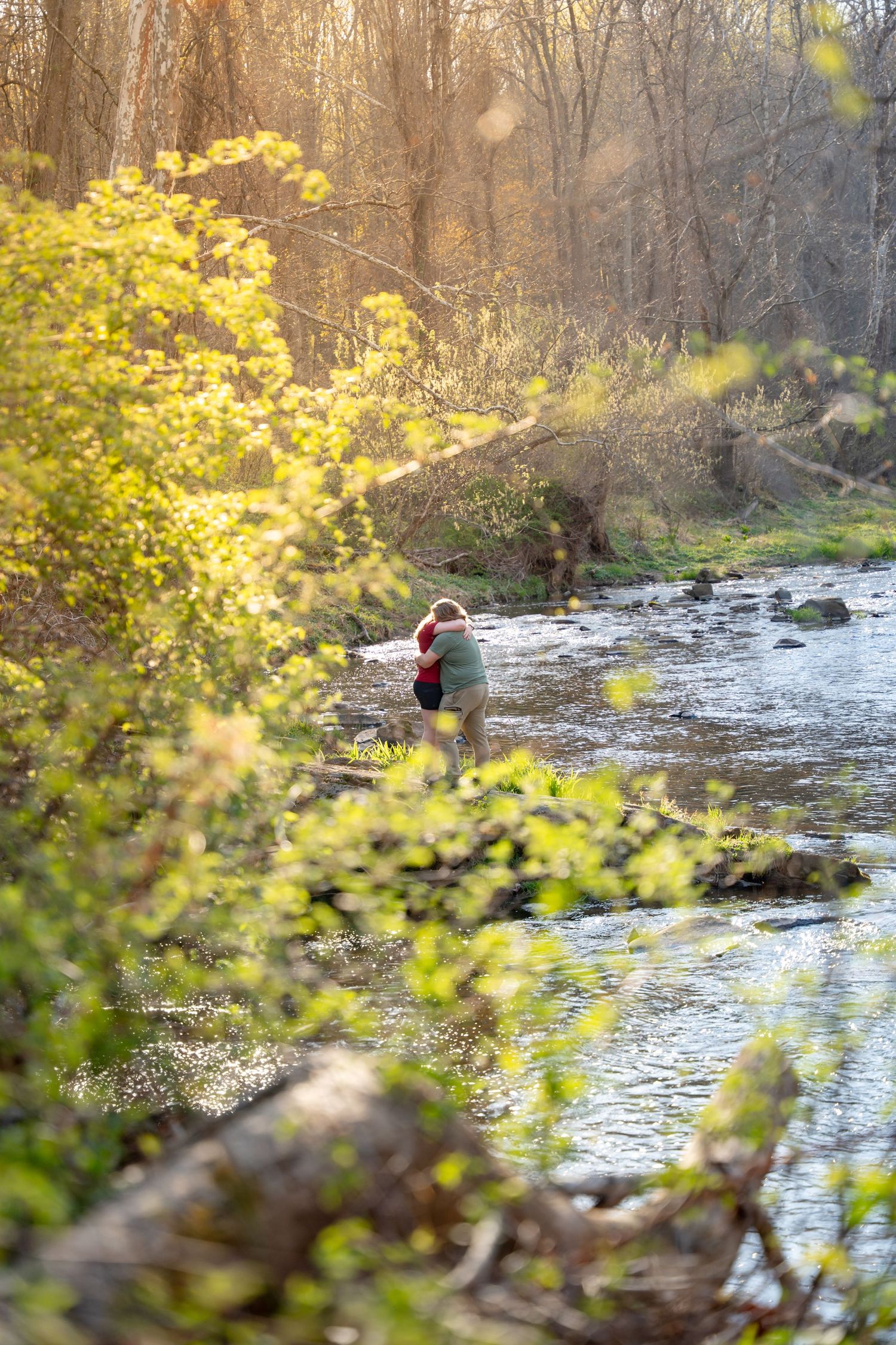 Two people embracing by a rocky stream in a sun-drenched forest with golden autumn foliage. during a proposal In Jerusalem mill in Fallston