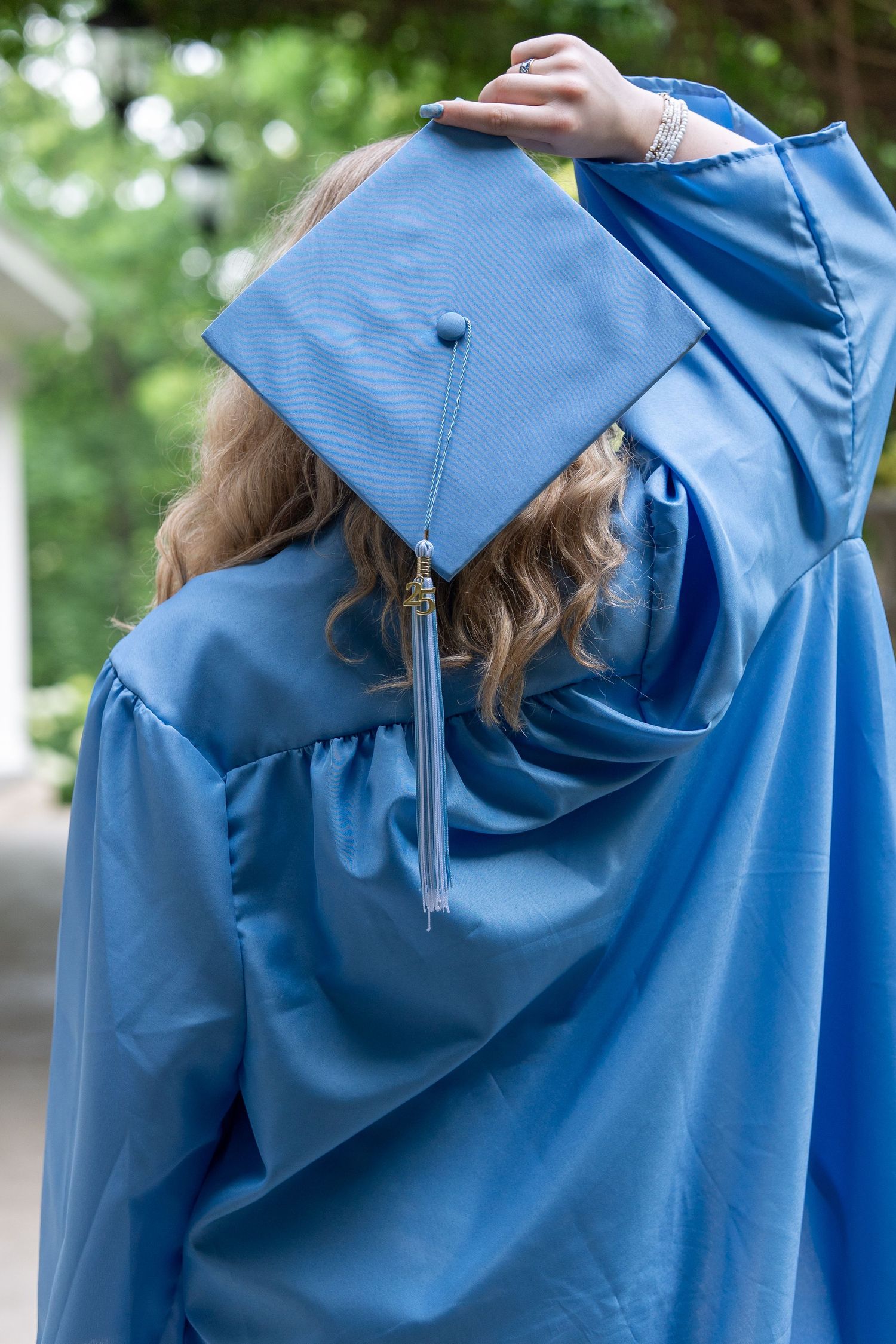 Back view of a female graduate in light blue cap and gown adjusting her mortarboard hat outdoors. In Bel Air Maryland
