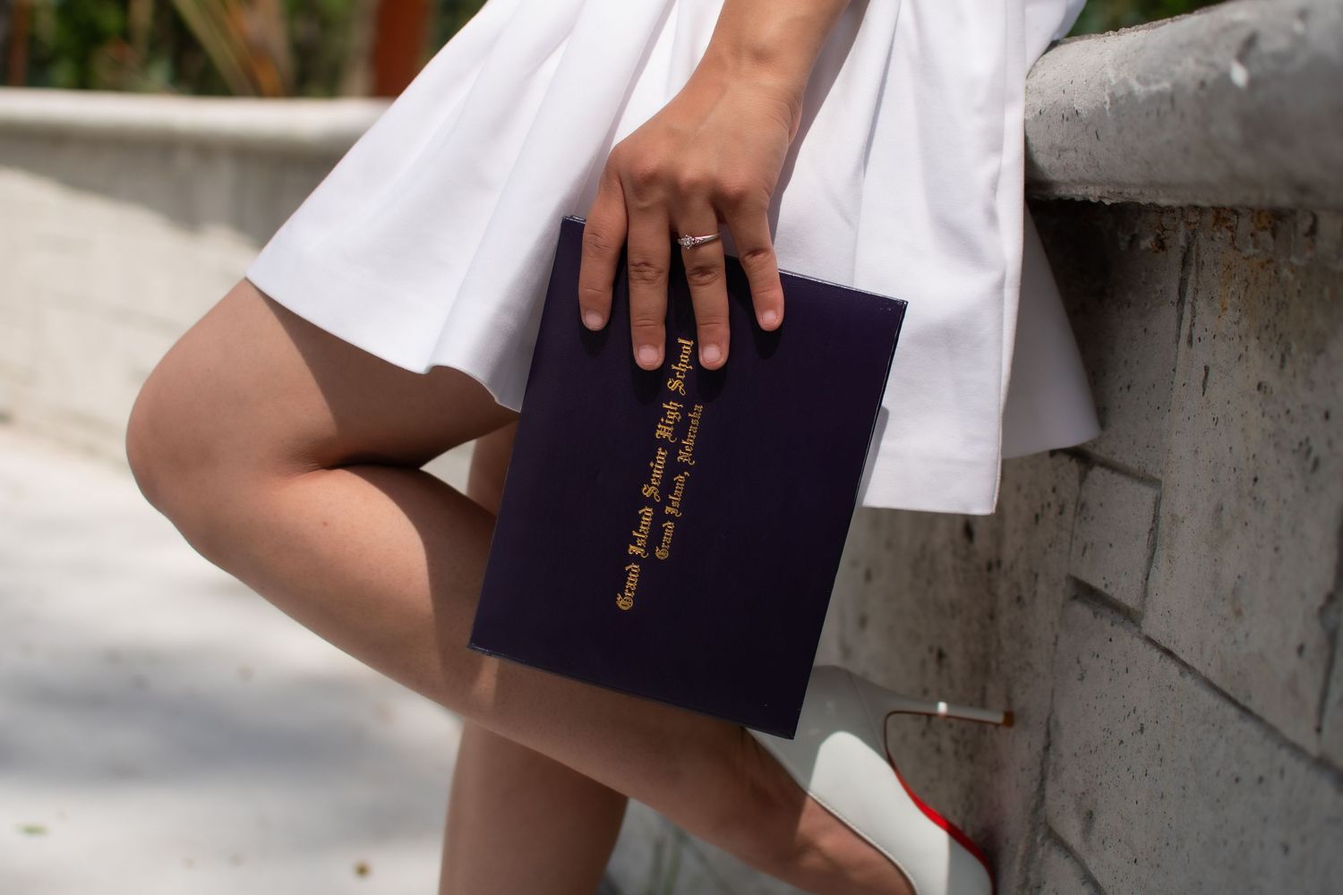 Girl in white dress and red heels (with red bottoms) leaning on concrete with one leg kicked up and holding her diploma close to her.